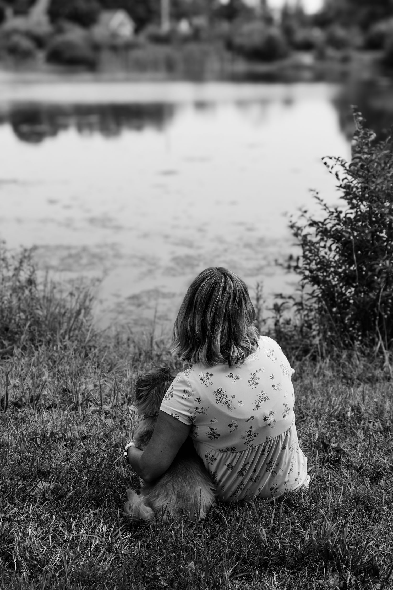 A person in a floral dress sits alone on grassy riverbank in black and white photograph.