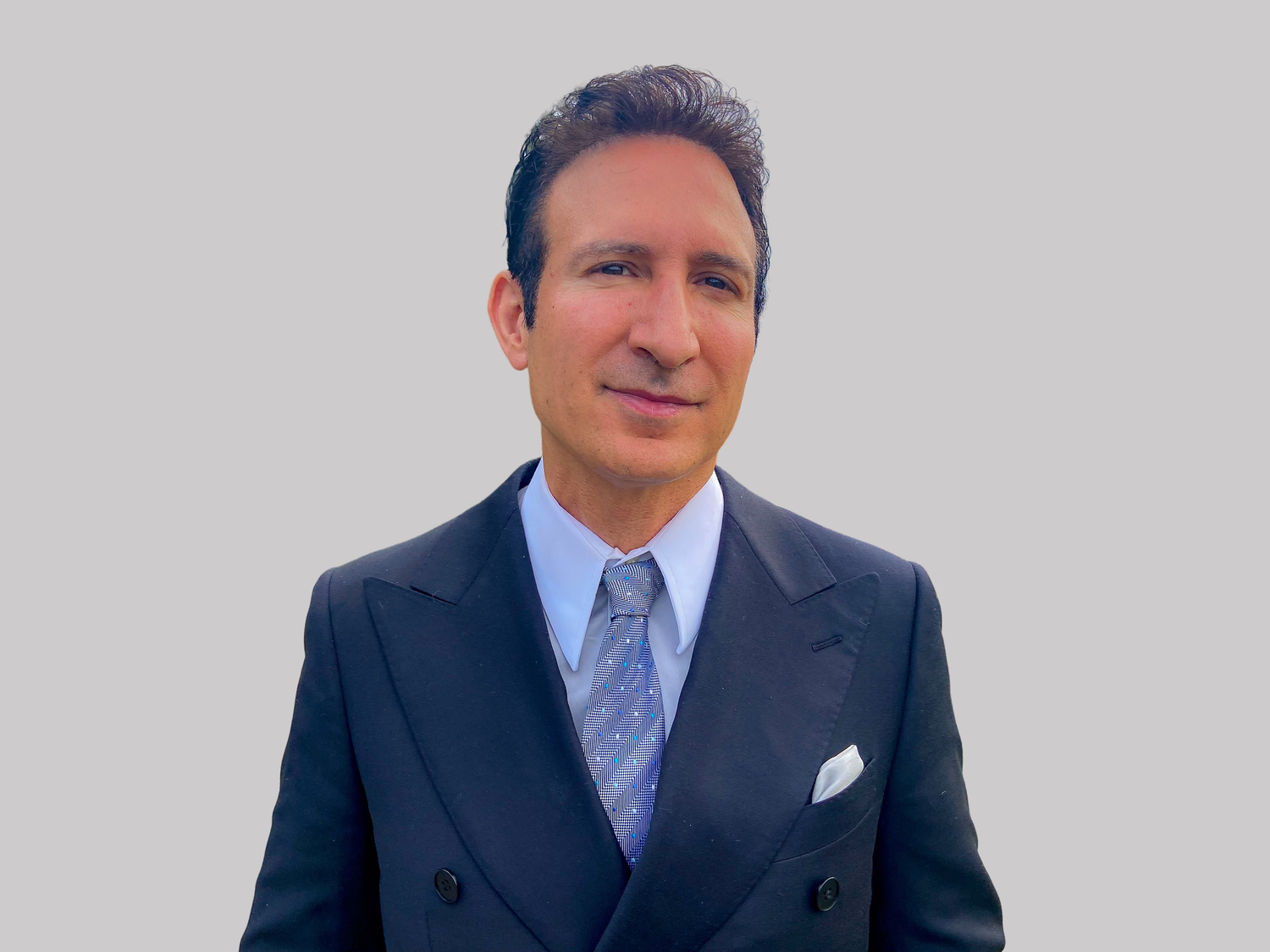 Professional portrait of someone wearing a navy double-breasted suit with light blue tie against neutral background.