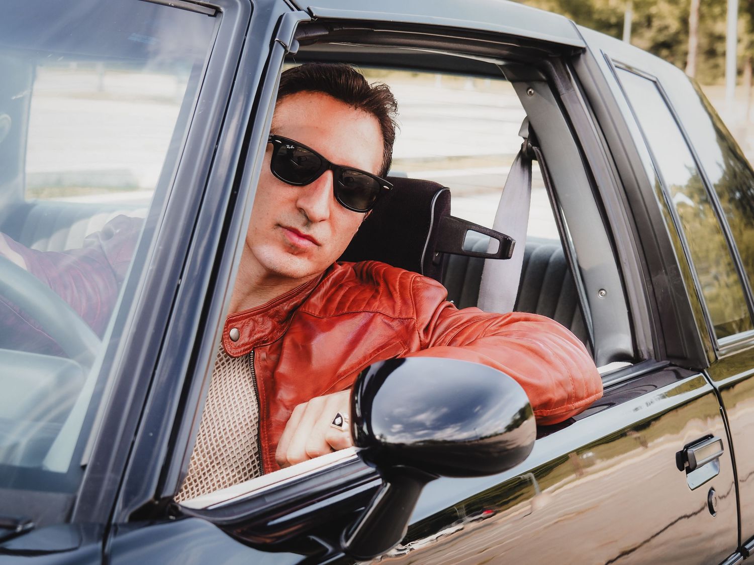 A person in sunglasses and red jacket sits in a car looking out the driver's side window on a sunny day.