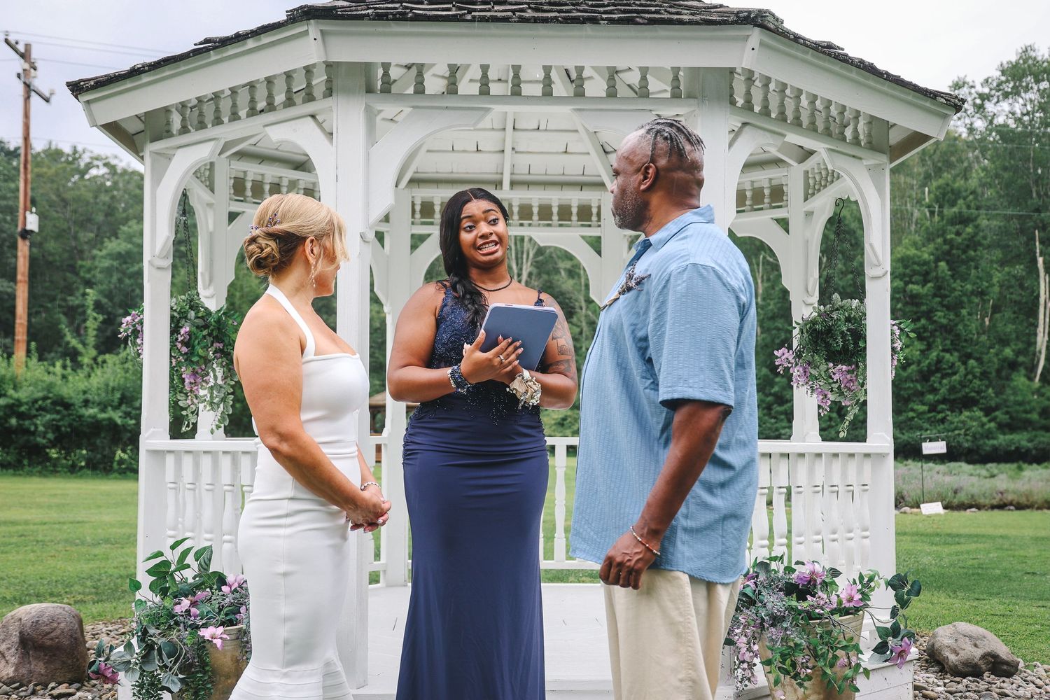 A wedding ceremony takes place at a white gazebo surrounded by lush greenery and gardens.