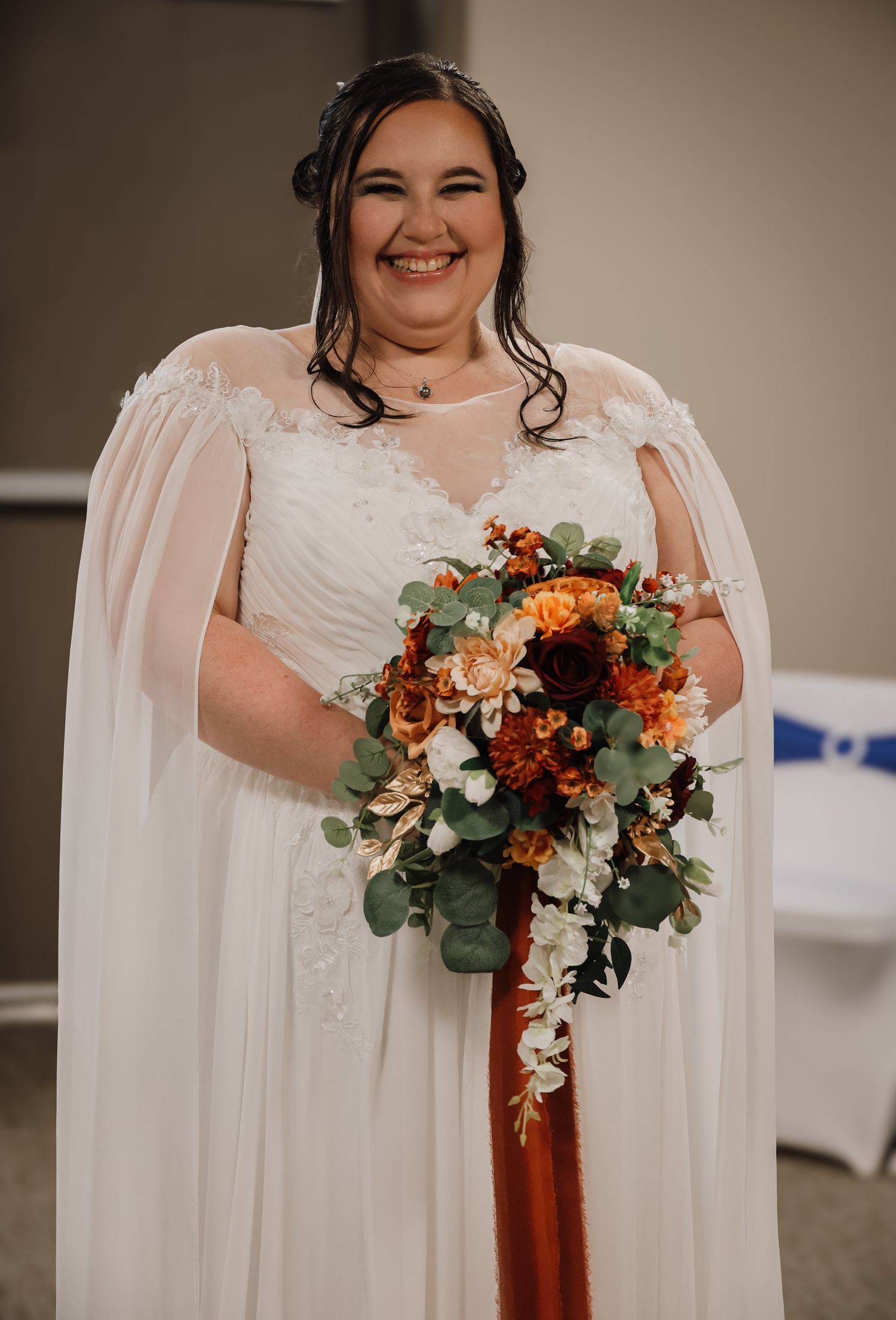 Bride in white lace dress holds vibrant autumn-colored bouquet with orange and burgundy flowers against white backdrop.