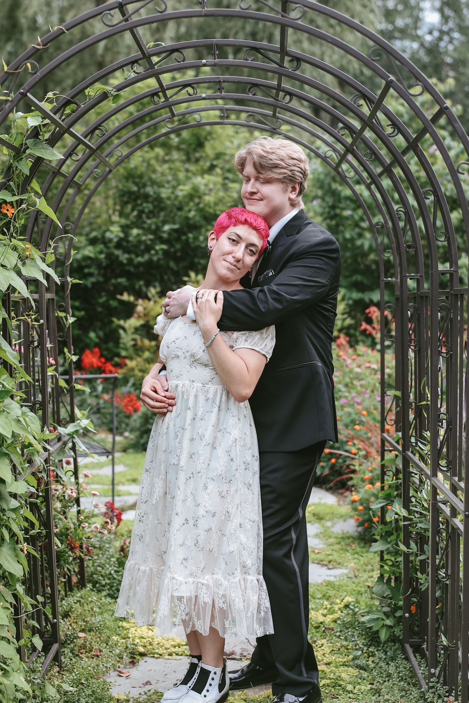 A couple embraces under a garden archway, with the person in white wearing a lace dress and sneakers.