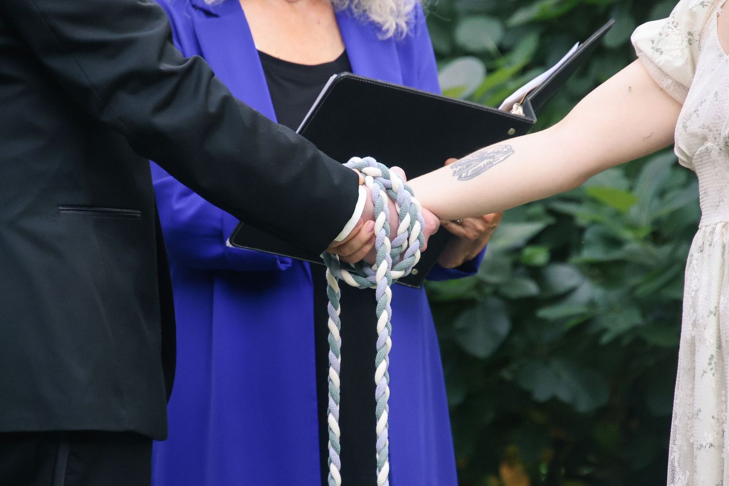 Close-up of hands exchanging wedding rings during an outdoor ceremony with purple and black attire.