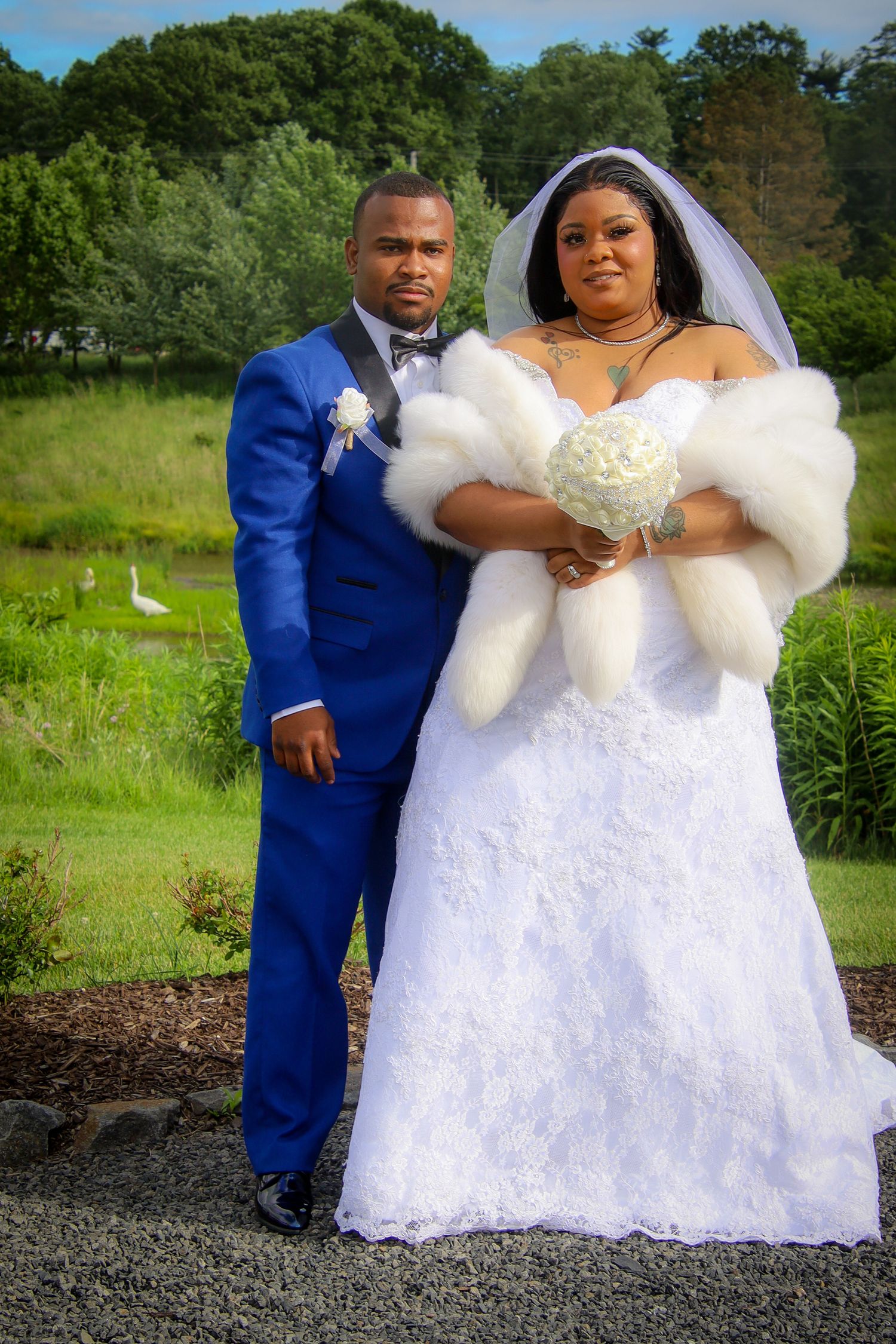 A wedding couple poses together outdoors in formal attire with the bride wearing a white lace gown and veil.