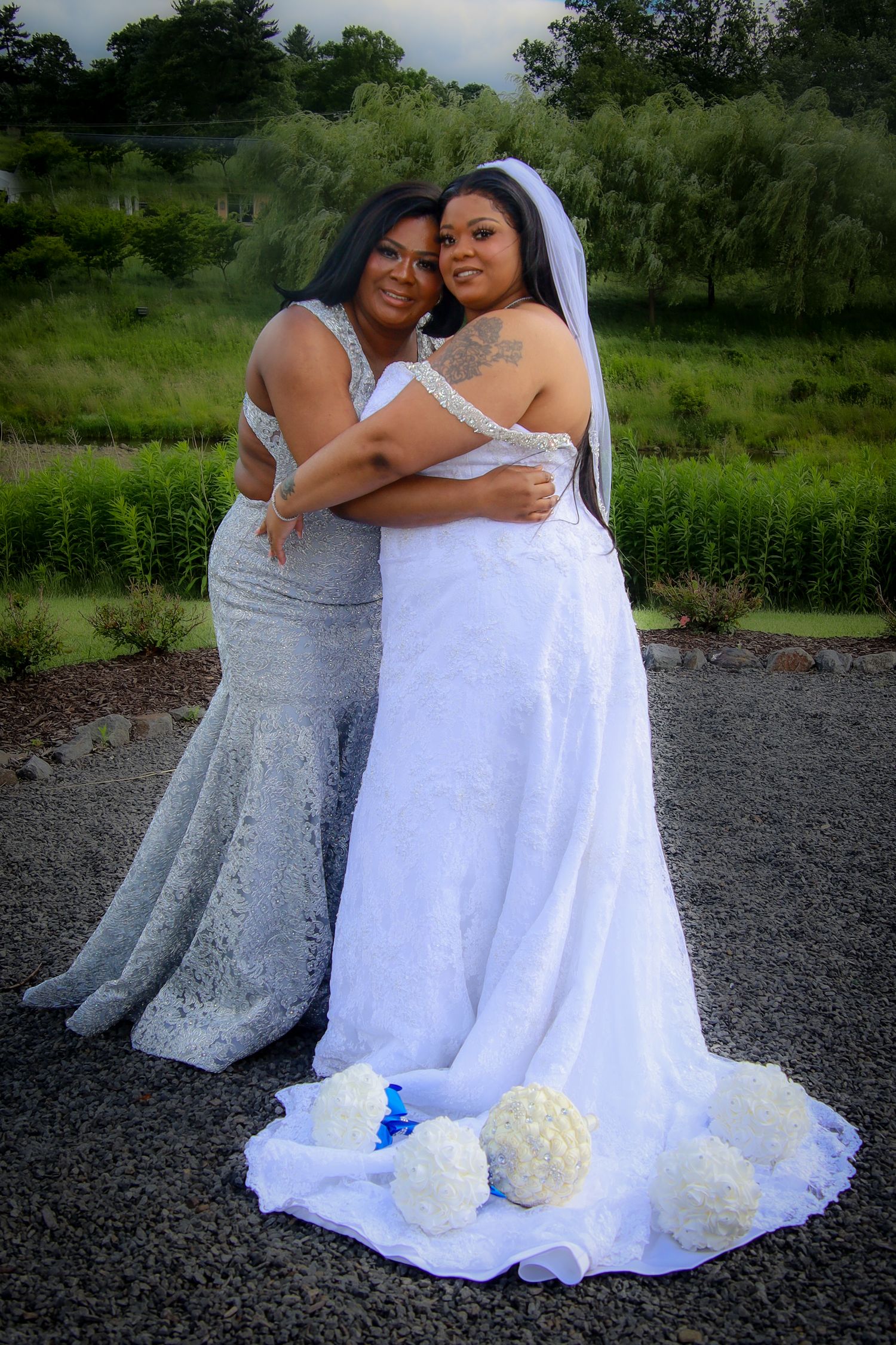 Two people in white wedding dresses embrace while sitting together outdoors on rocky ground.