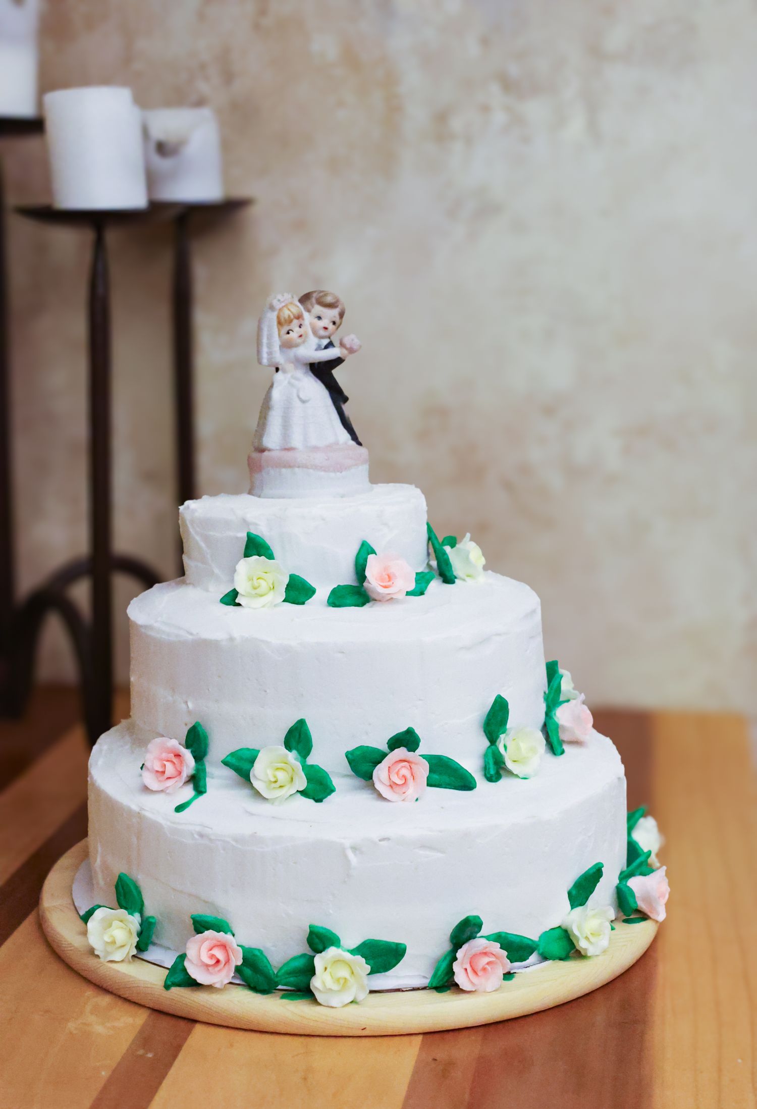 A three-tiered white wedding cake decorated with pink and cream roses, green leaves, and a bride and groom topper.
