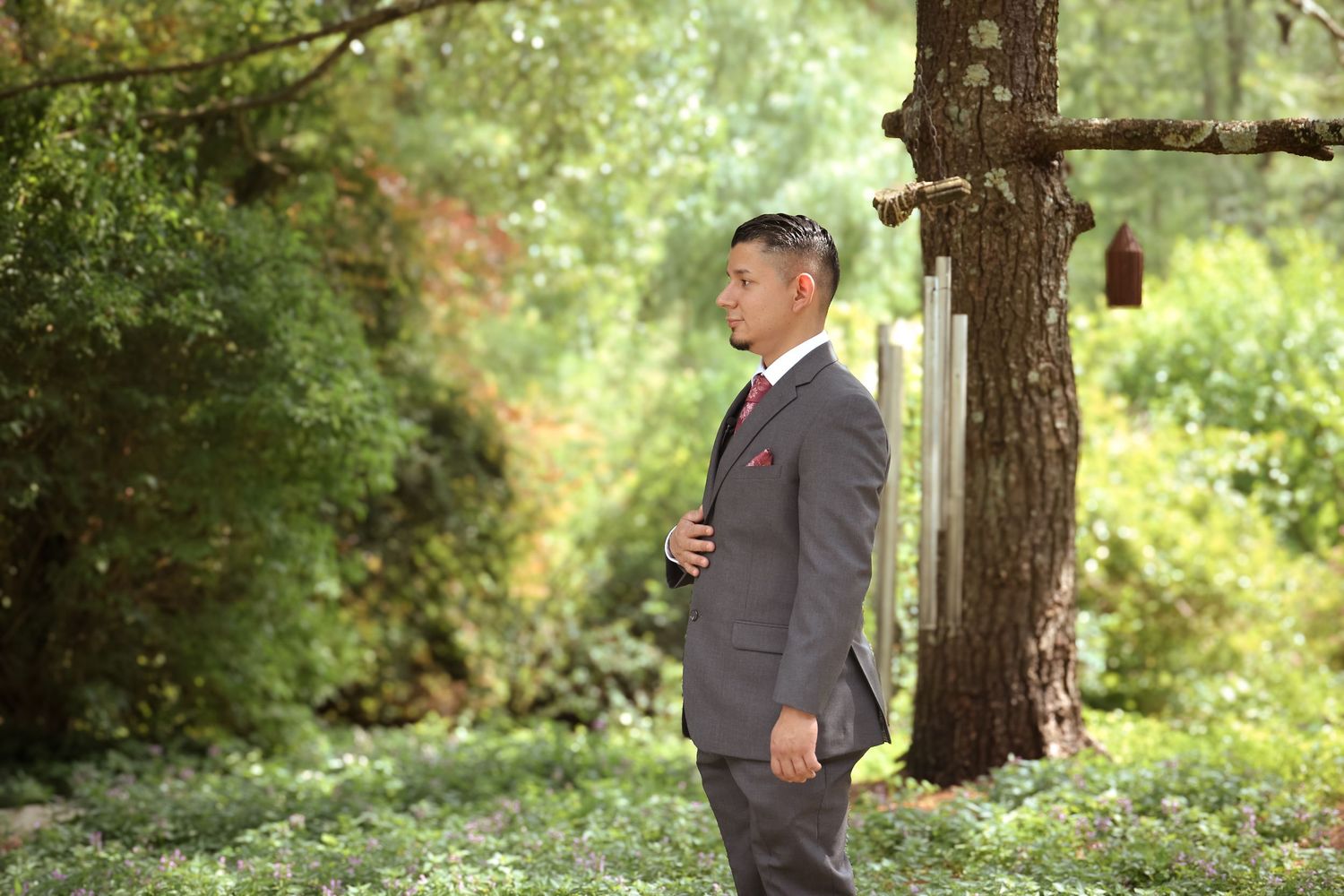 A couple in formal attire stands together in a scenic wooded outdoor setting during a wedding photoshoot.