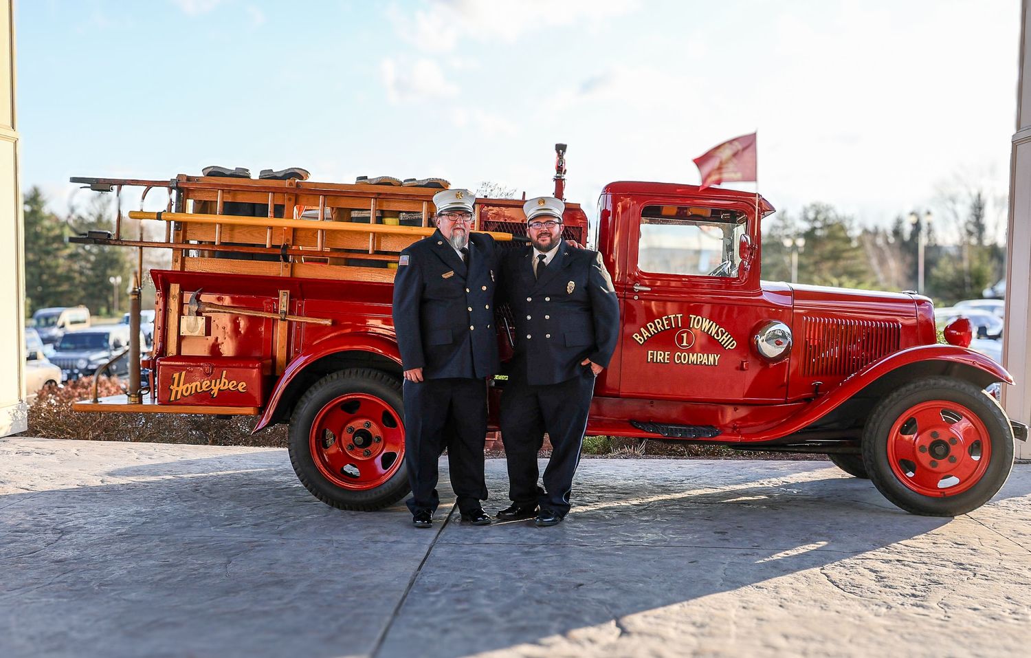 Two firefighters in uniform standing in front of a vintage red fire truck.