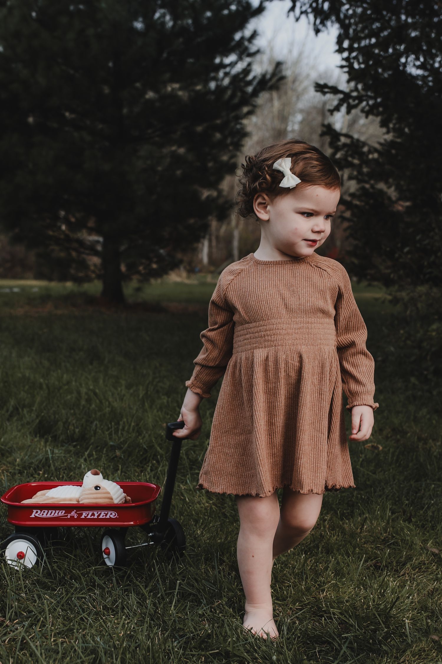 Young child in brown dress standing barefoot next to red Radio Flyer wagon in grassy field.