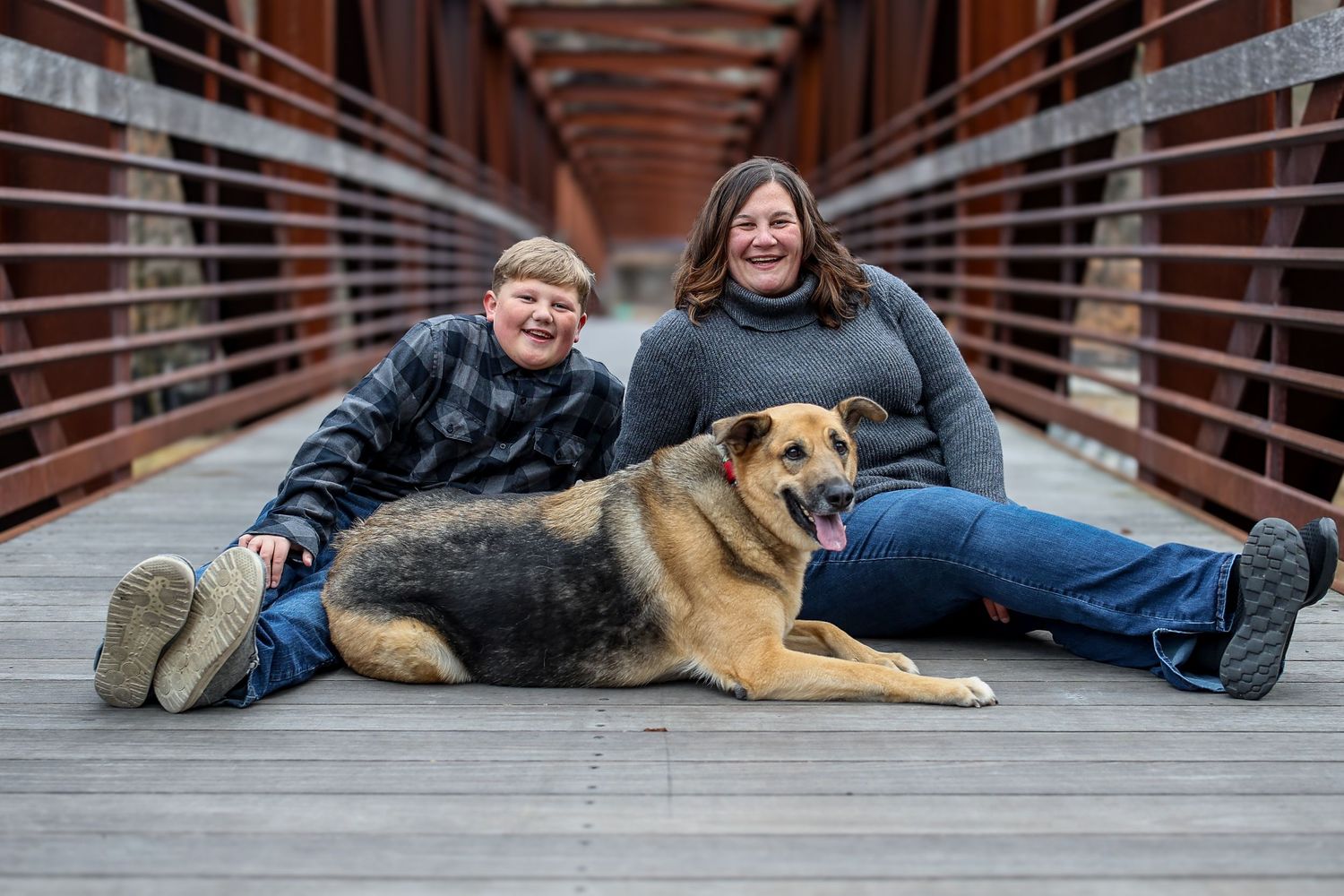 A person in jeans and boots lies on wooden deck boards next to a relaxed German Shepherd dog.
