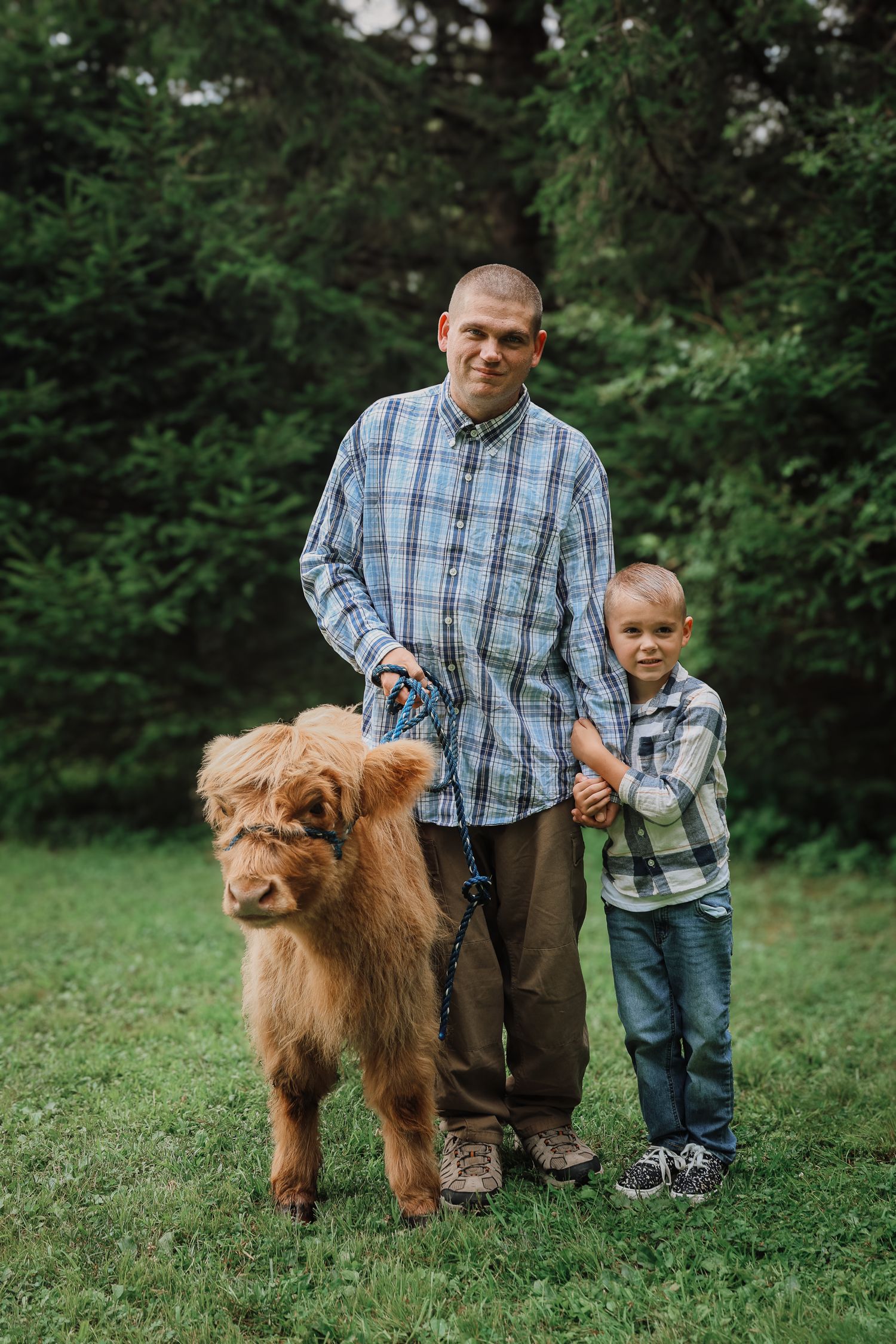 A brown fluffy dog stands on grass with people in a plaid shirt and casual attire outdoors.