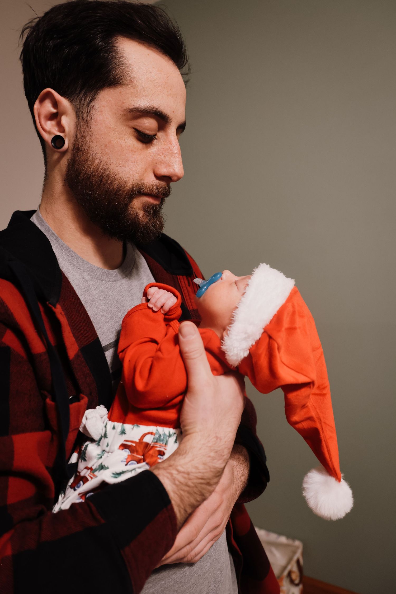A tender holiday moment shows someone in a red plaid shirt holding a sleeping baby wearing an orange outfit and Santa hat.