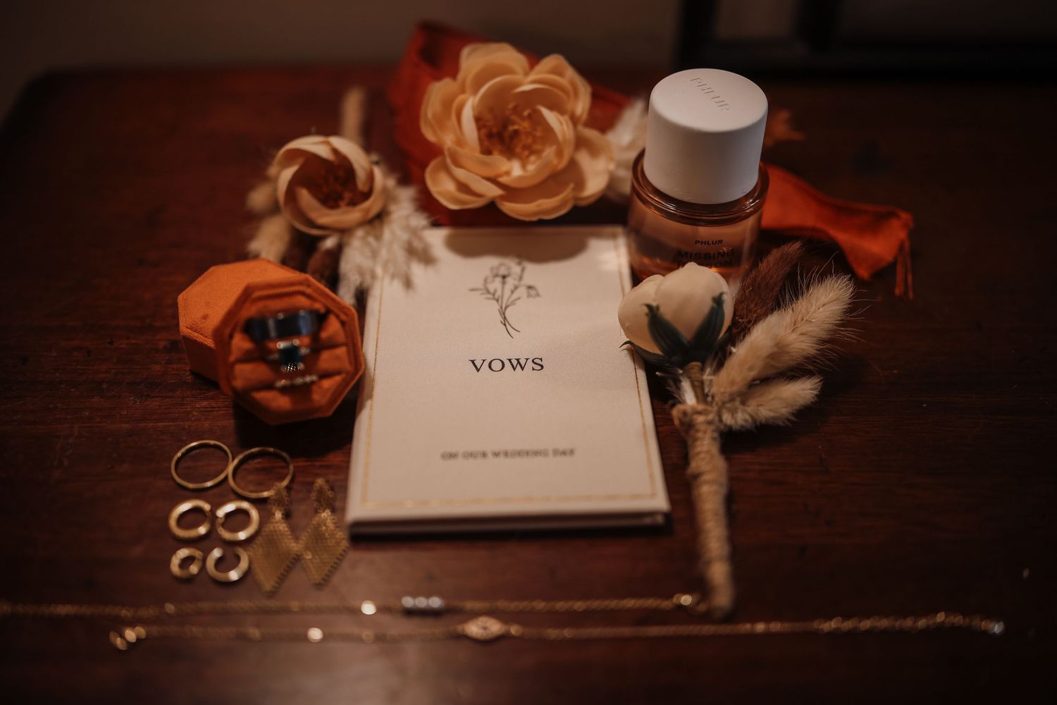Wedding rings and decorative details arranged on dark wooden surface with orange floral accents and paper elements.