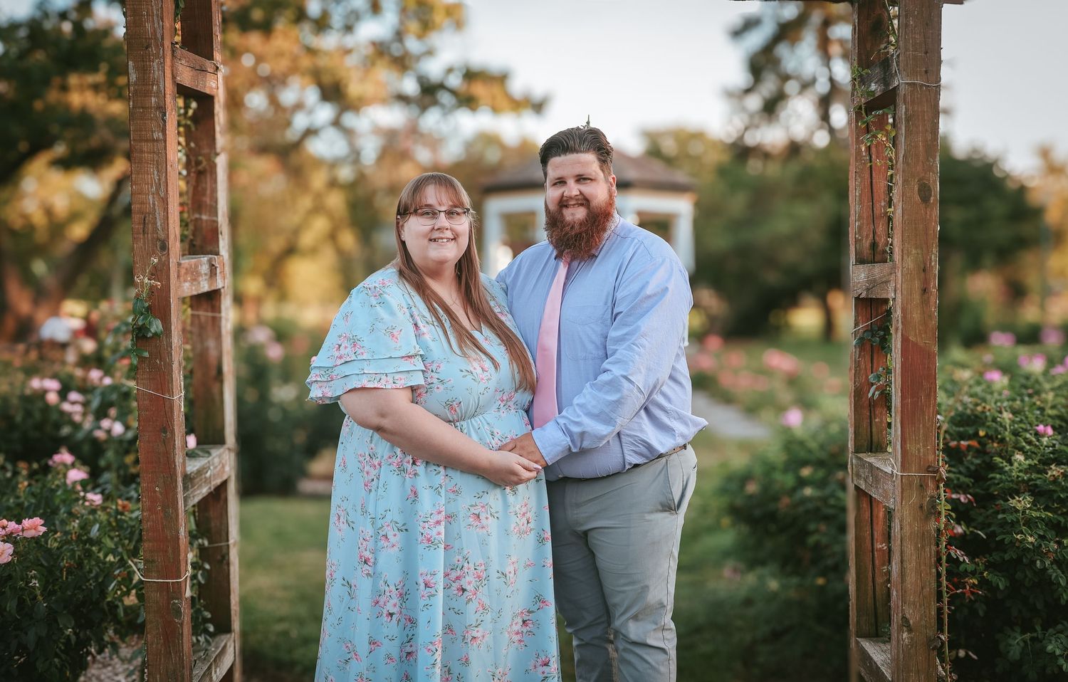 A couple stands together under a rustic wooden archway in a garden setting during golden hour.