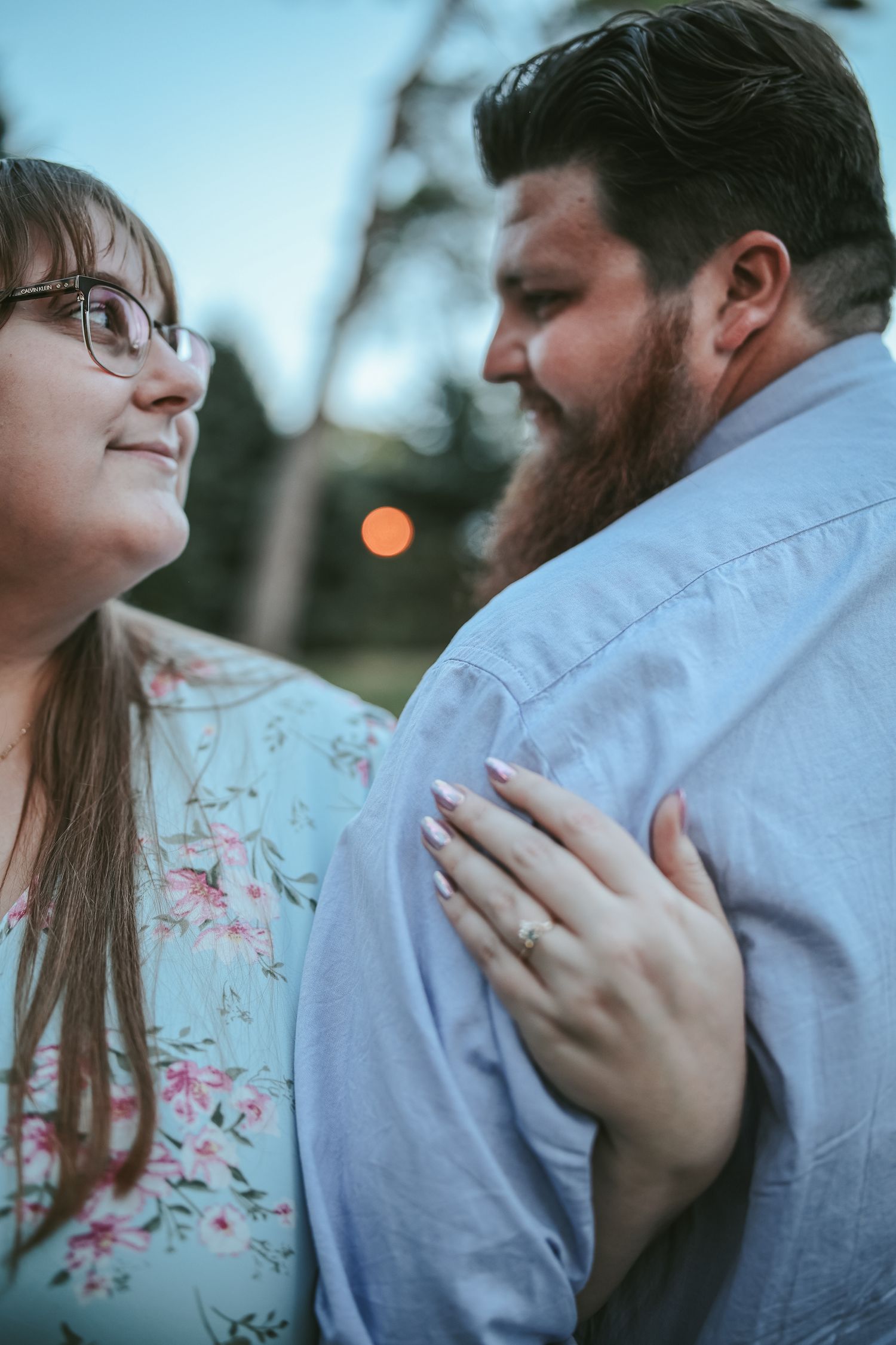 A romantic couple in a floral dress and blue shirt embrace outdoors during a sunset photoshoot.