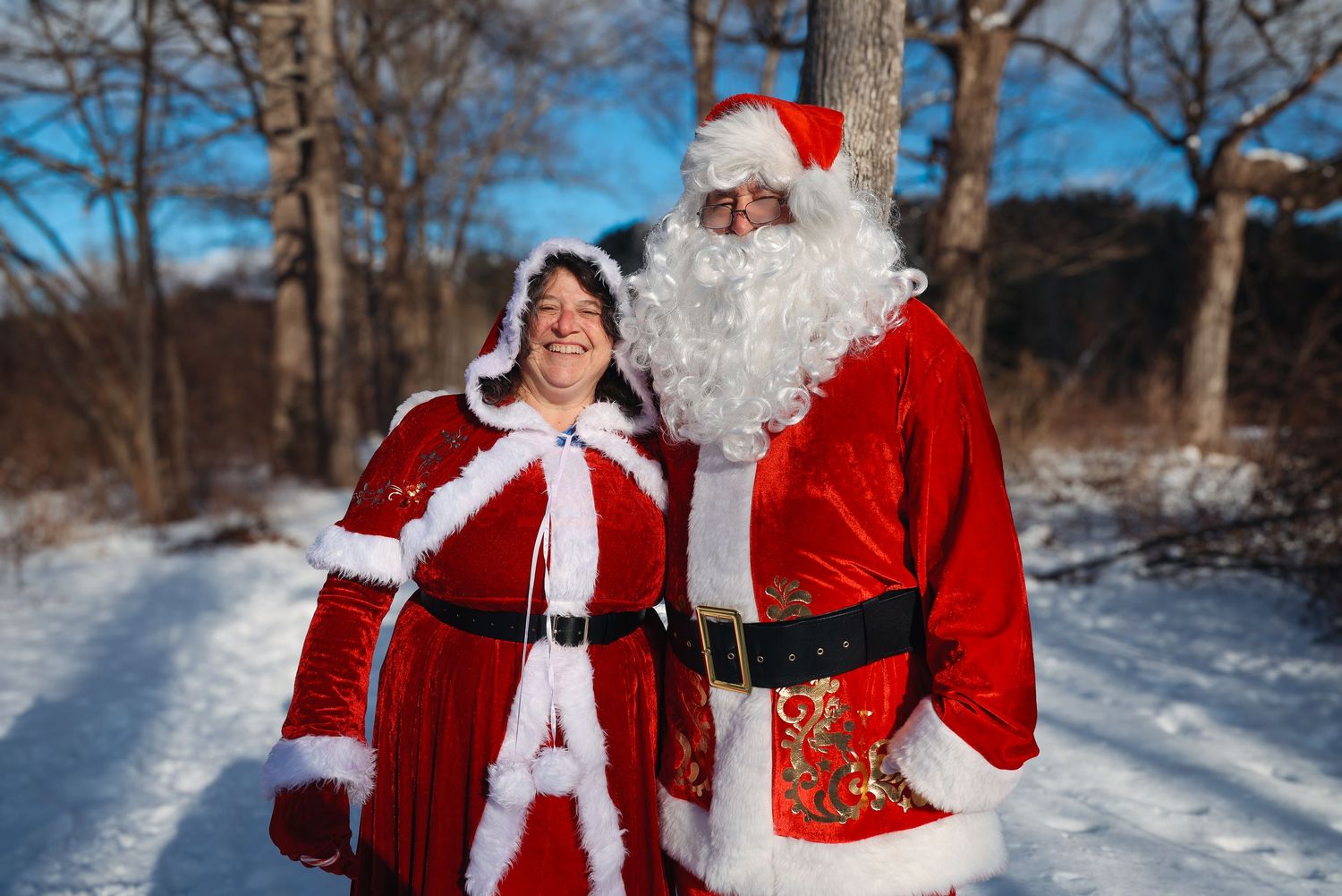 Two festive figures in red and white Santa Claus costumes stand together on a snowy winter path surrounded by bare trees.