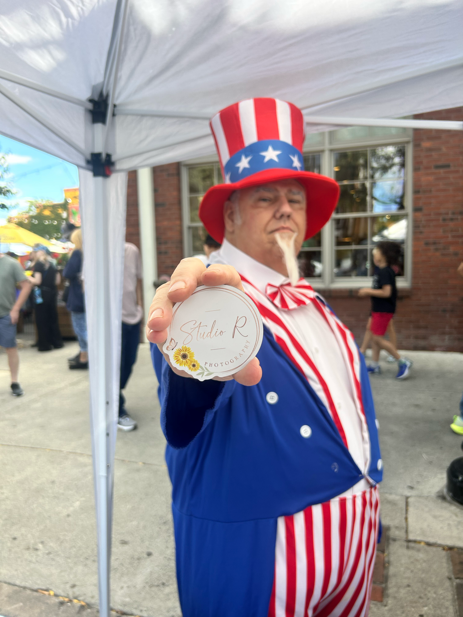 A performer in a patriotic Uncle Sam costume and stars-and-stripes outfit waves while standing under a white tent canopy.