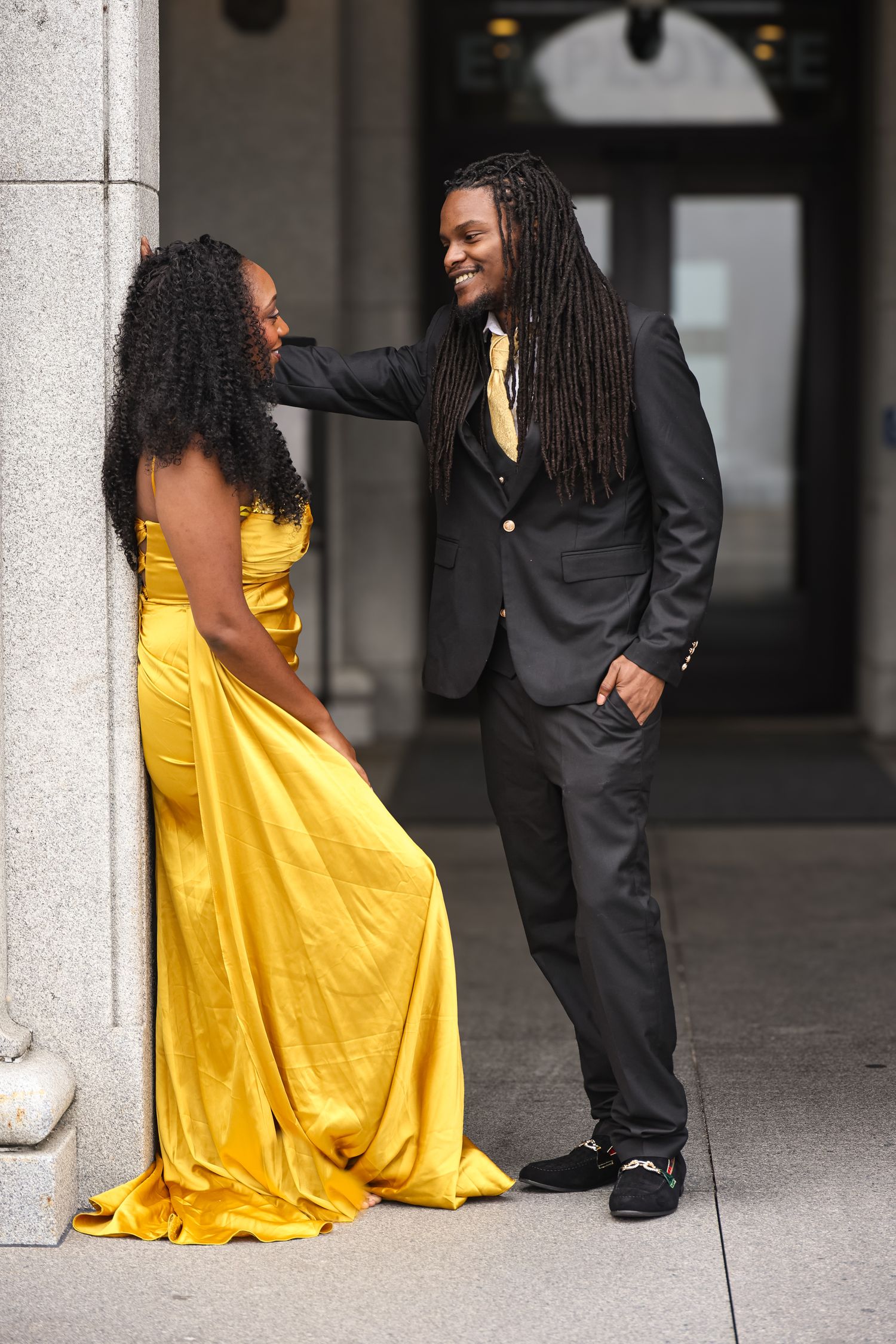 A couple poses together outdoors, with one wearing a bright yellow formal gown and the other in a black suit.