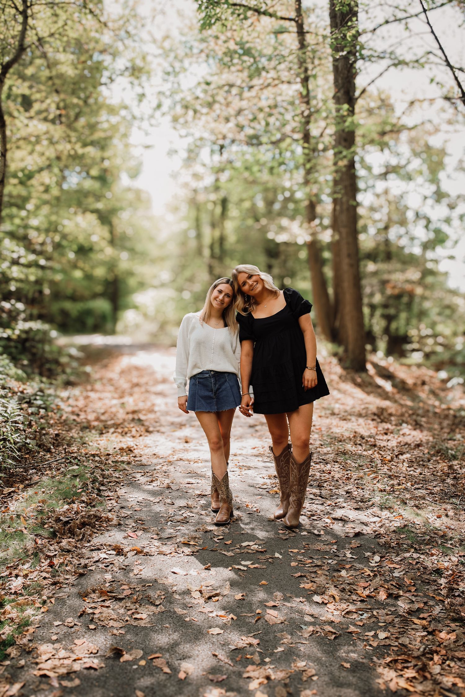 Two people walk together on a forest trail covered in fallen autumn leaves in soft natural lighting.