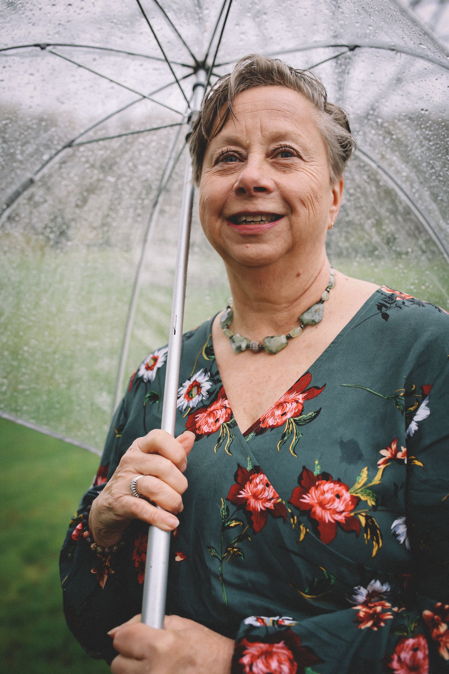 A person in a floral dress holds a clear umbrella while smiling on a rainy day outdoors.
