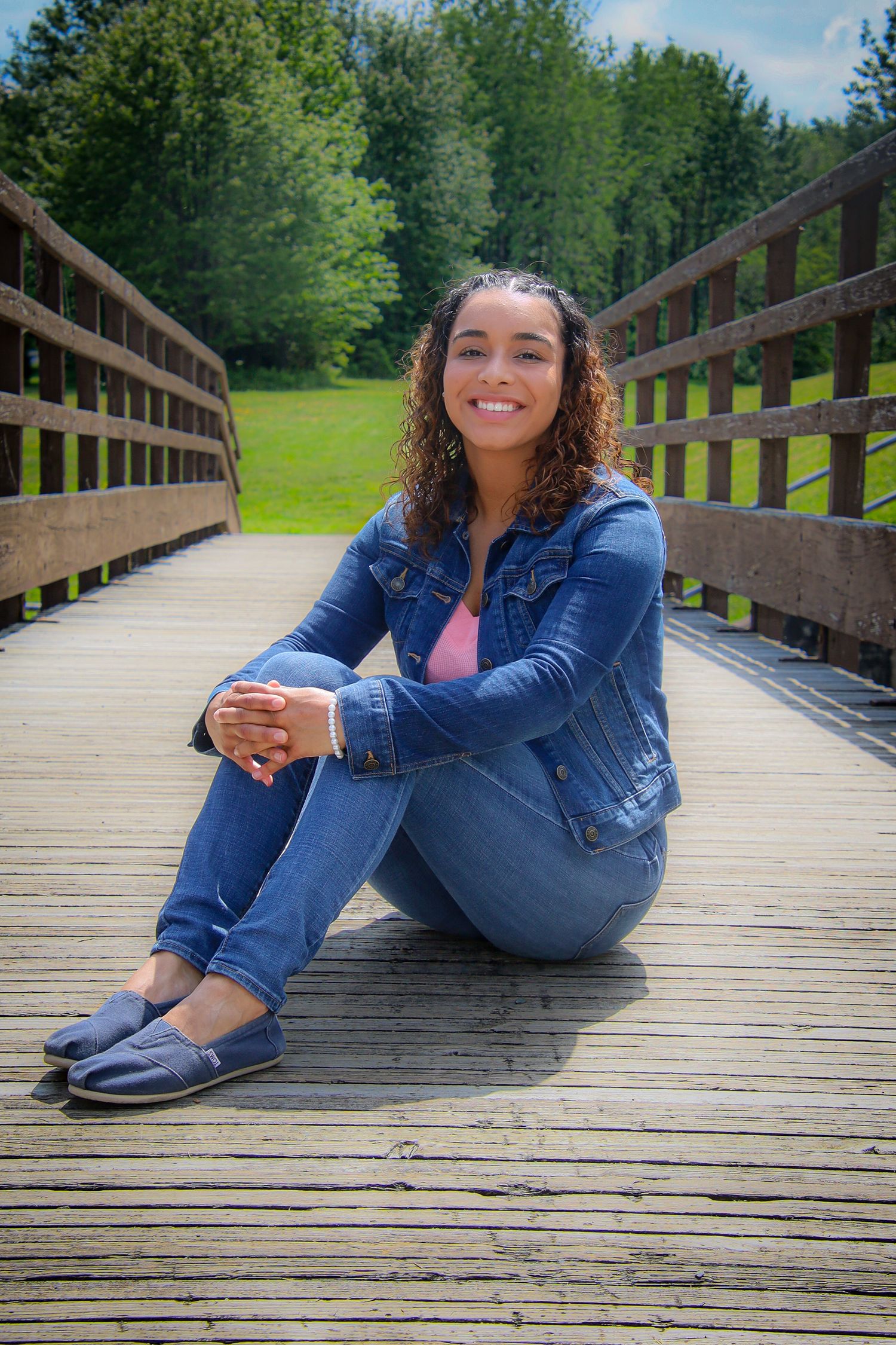 Person in denim outfit sits casually on wooden boardwalk surrounded by lush green forest scenery.