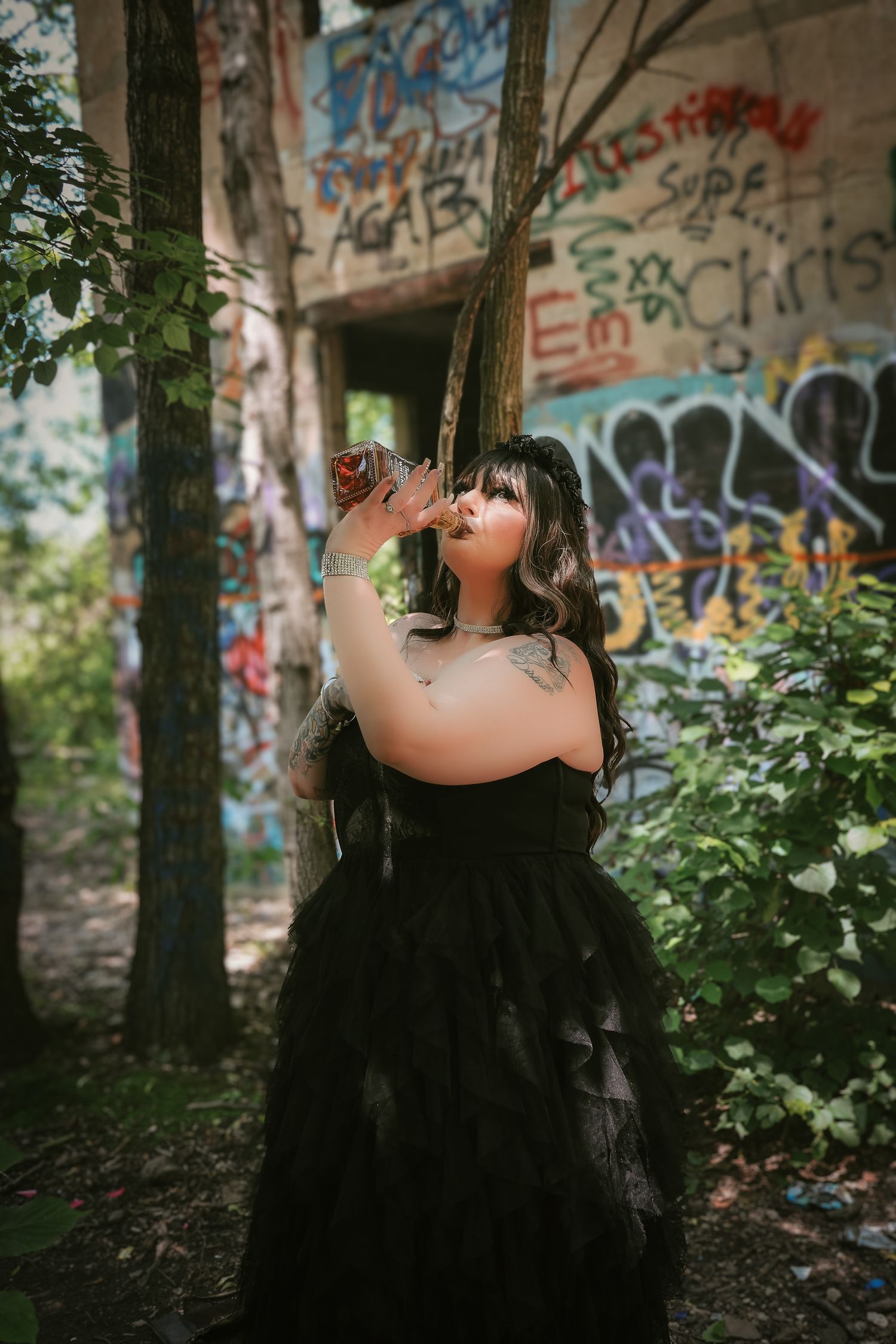 A person in a black dress poses near graffiti-covered walls in a wooded area, creating a dramatic artistic scene.