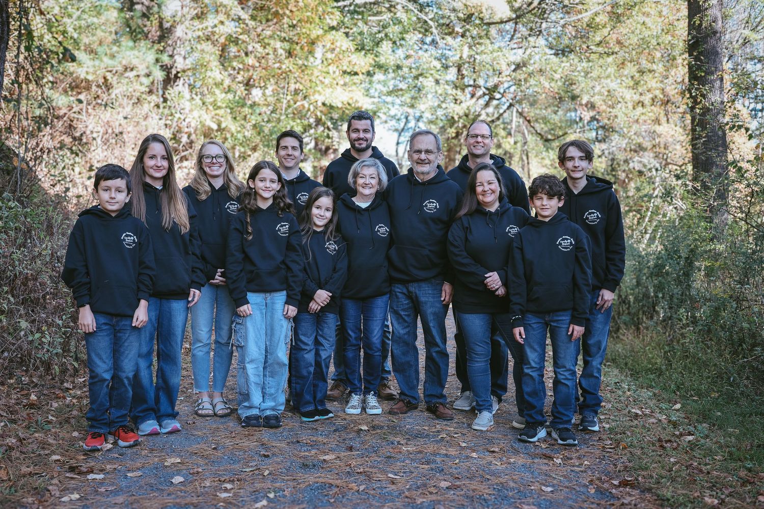 A group of people wearing dark clothing pose together outdoors in an autumn setting with fallen leaves.