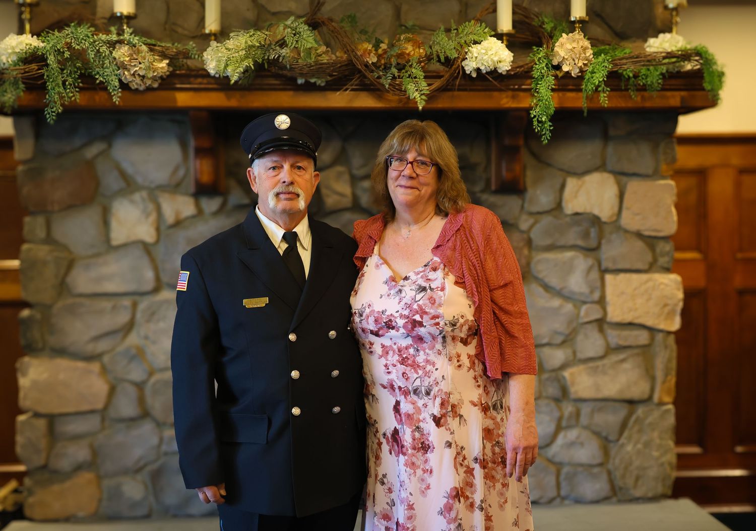 A couple stands together in front of a stone wall at an indoor venue with decorative greenery overhead.
