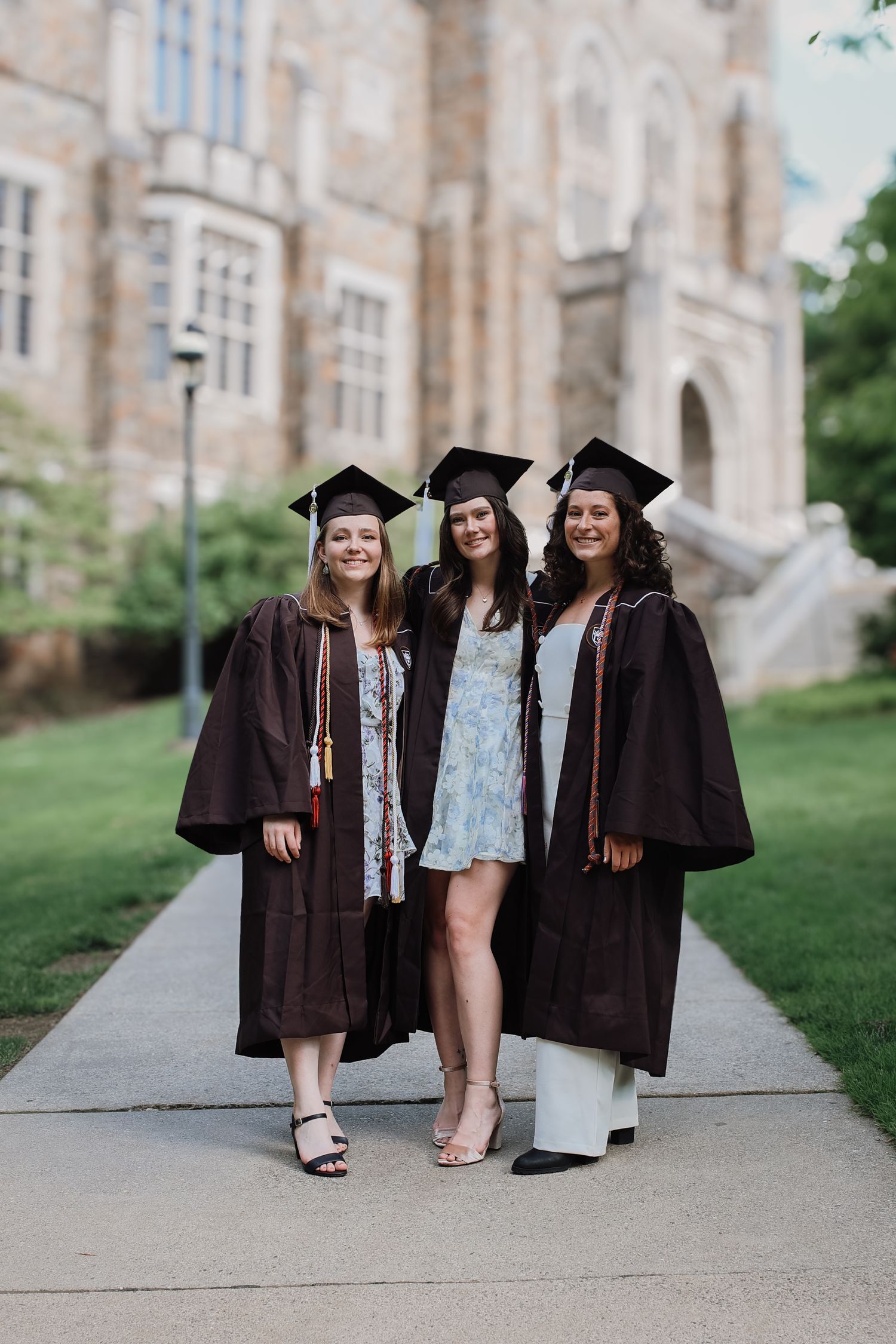 Three graduates in black caps and gowns pose together on a stone walkway near a university building.