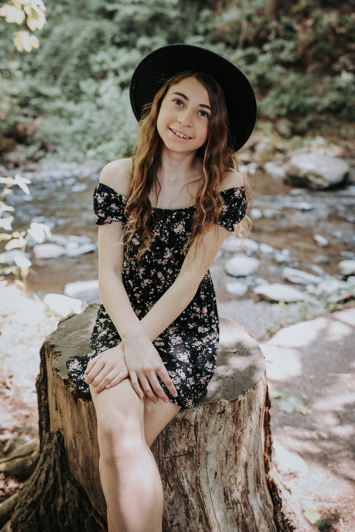 A person in a black floral dress and wide-brimmed hat poses on a tree stump in a natural outdoor setting.