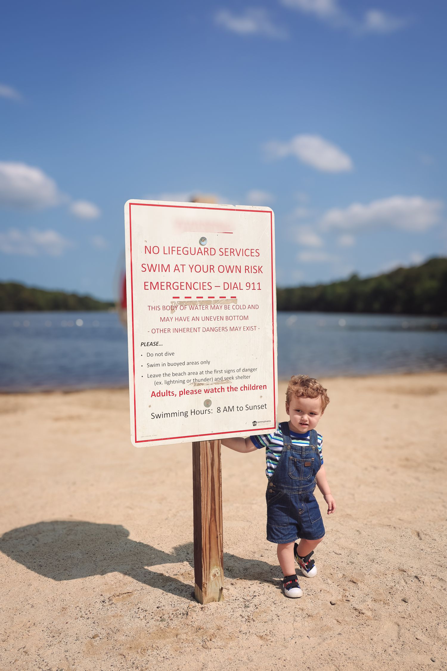 A warning sign stands on a sandy beach next to a small child in denim overalls by the water on a sunny day.