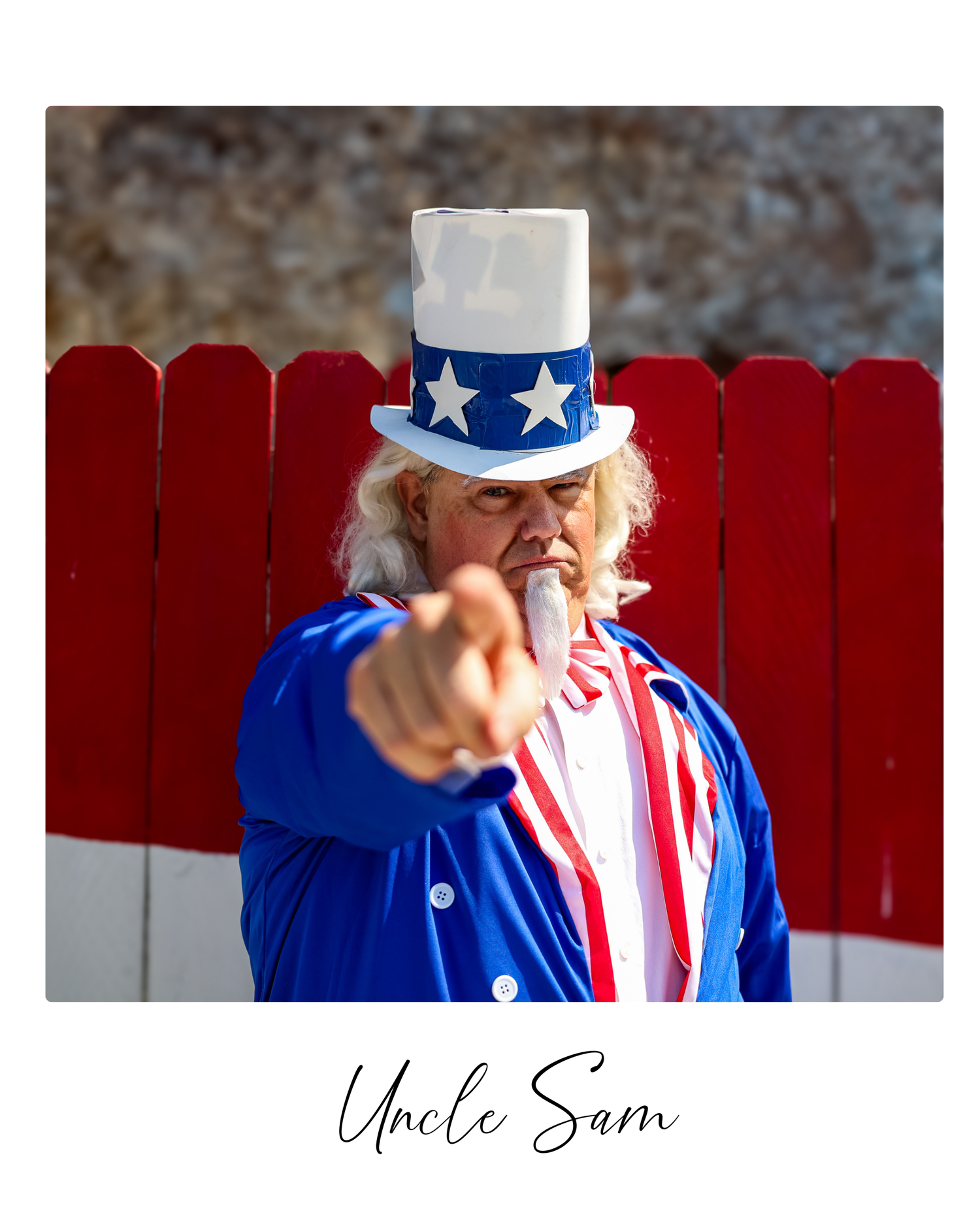 A patriotic figure in Uncle Sam costume with stars and stripes attire poses against a red fence in an outdoor setting.