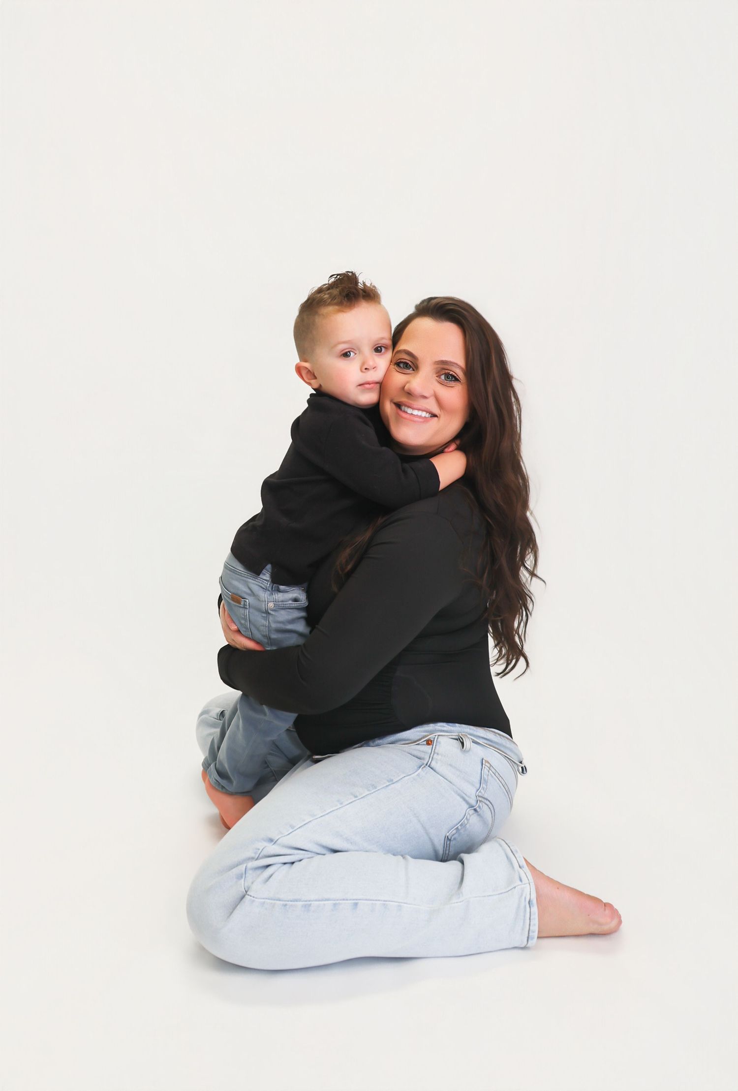 A woman with long dark hair sits on the floor hugging a young boy, both wearing black tops and jeans.