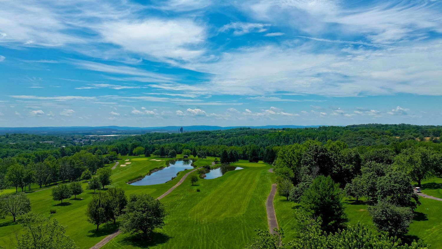 Aerial view of a lush green golf course with ponds, tree-lined fairways, and a blue sky with scattered clouds.