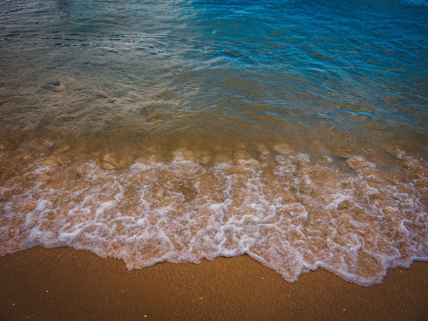 Turquoise ocean waves gently washing onto a sandy beach shore, creating white sea foam where water meets golden sand.