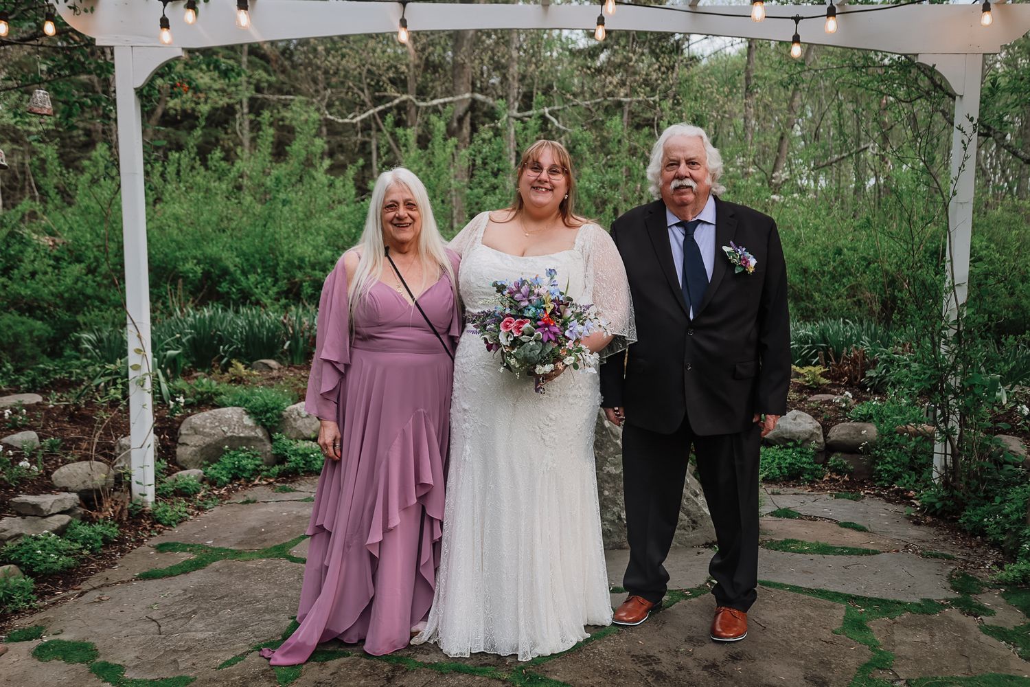 Bride in white gown poses with parents in garden wedding venue with string lights and stone path.