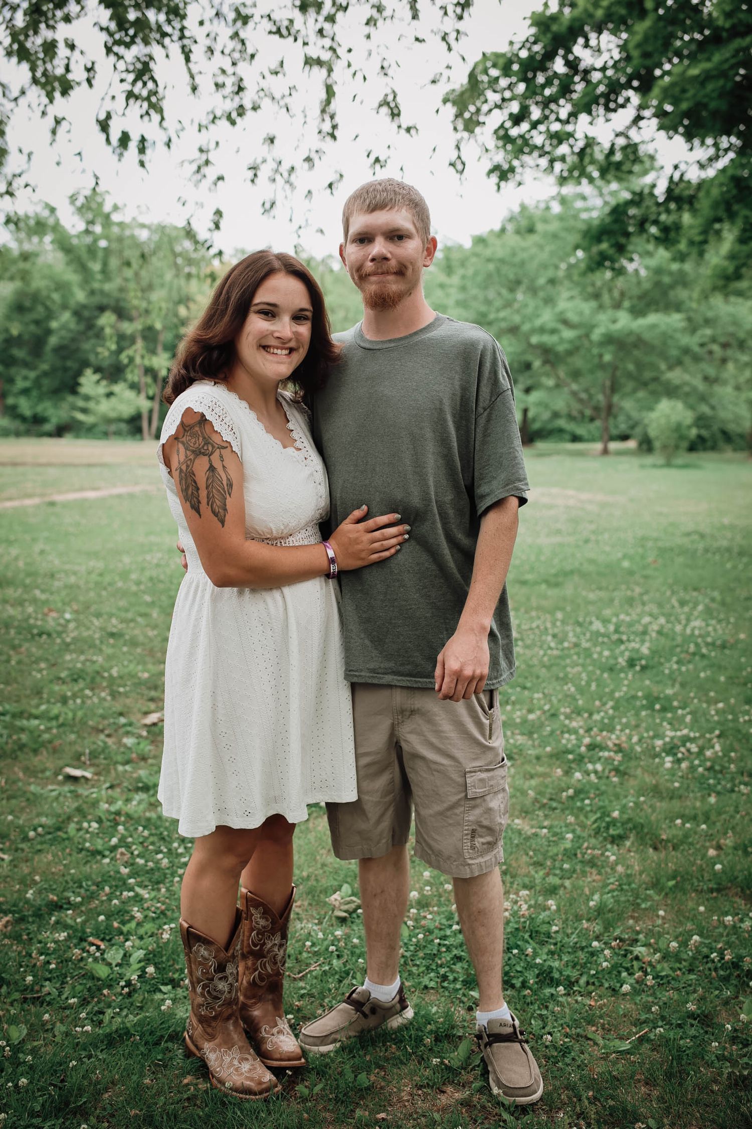 Couple posing outdoors on green grass, woman in white dress and cowboy boots, man in olive shirt and cargo shorts.