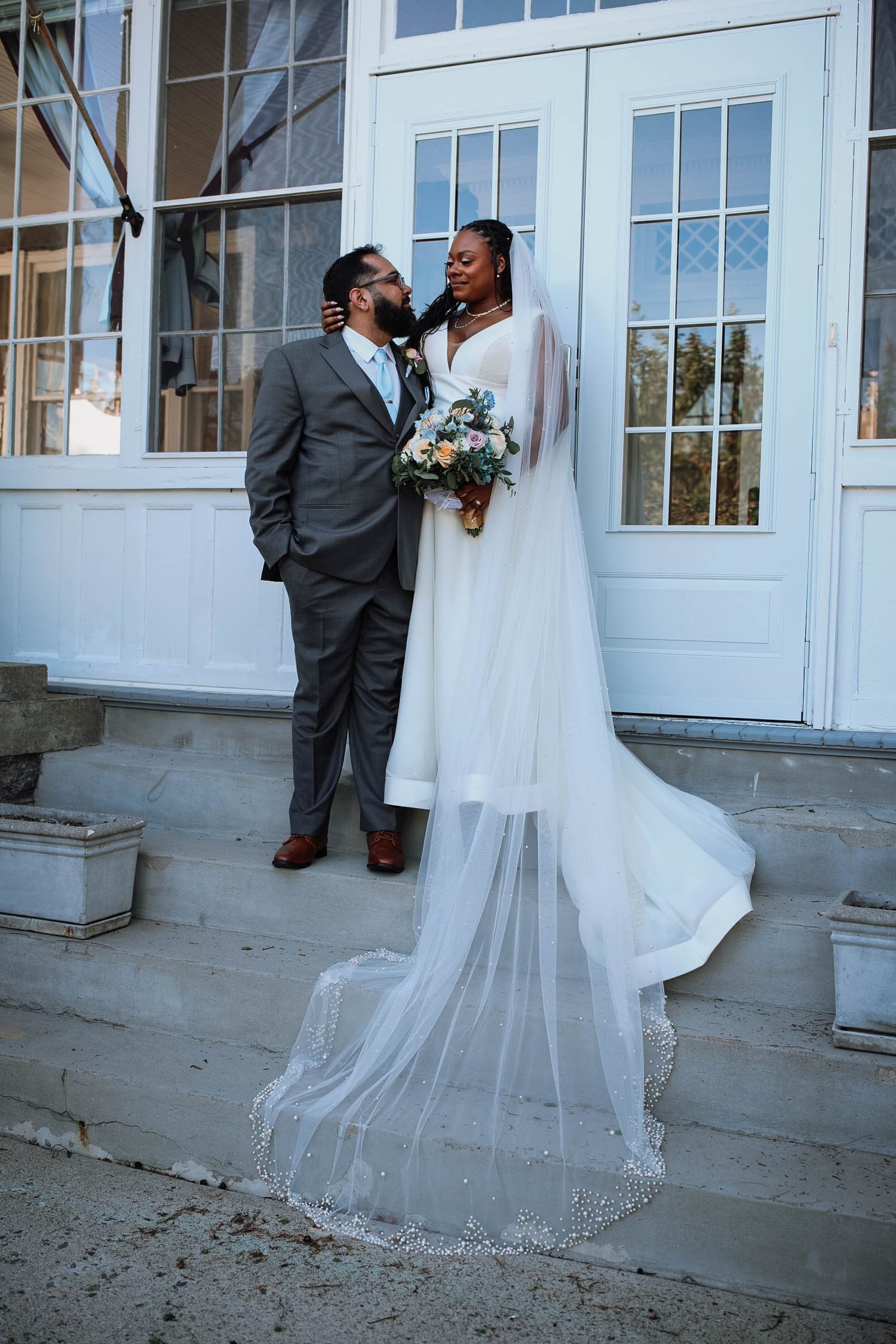 Bride in white gown with long veil and groom in gray suit share romantic moment on steps of elegant white building.