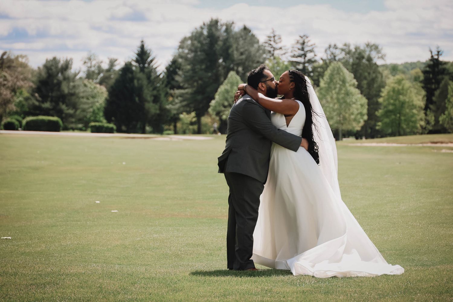 A bride and groom share a romantic embrace on a lush green lawn, surrounded by trees on their wedding day.