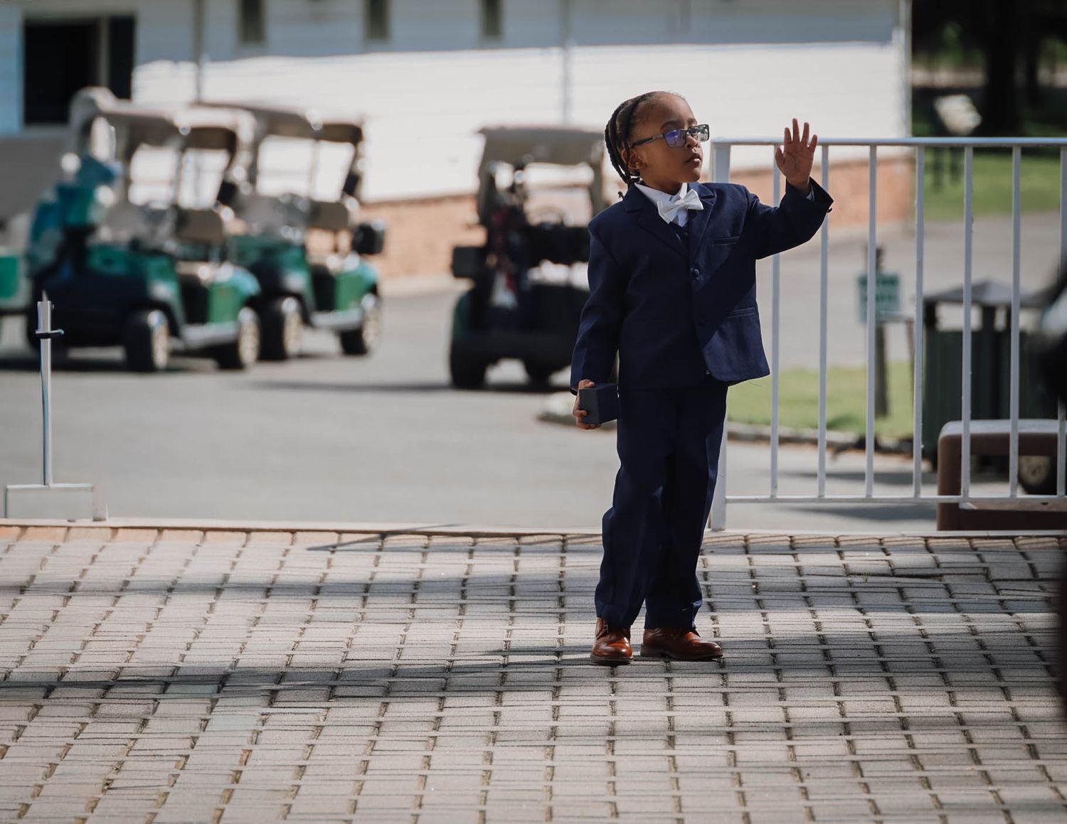 Young boy in navy suit waving outdoors near golf carts on a brick paved area.
