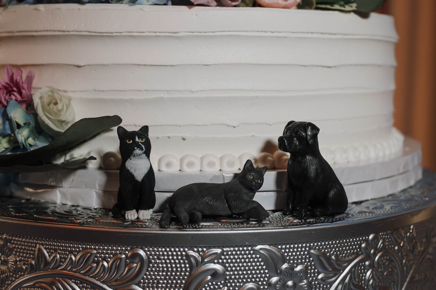 Three cat and dog figurines displayed on a silver tiered wedding cake stand.
