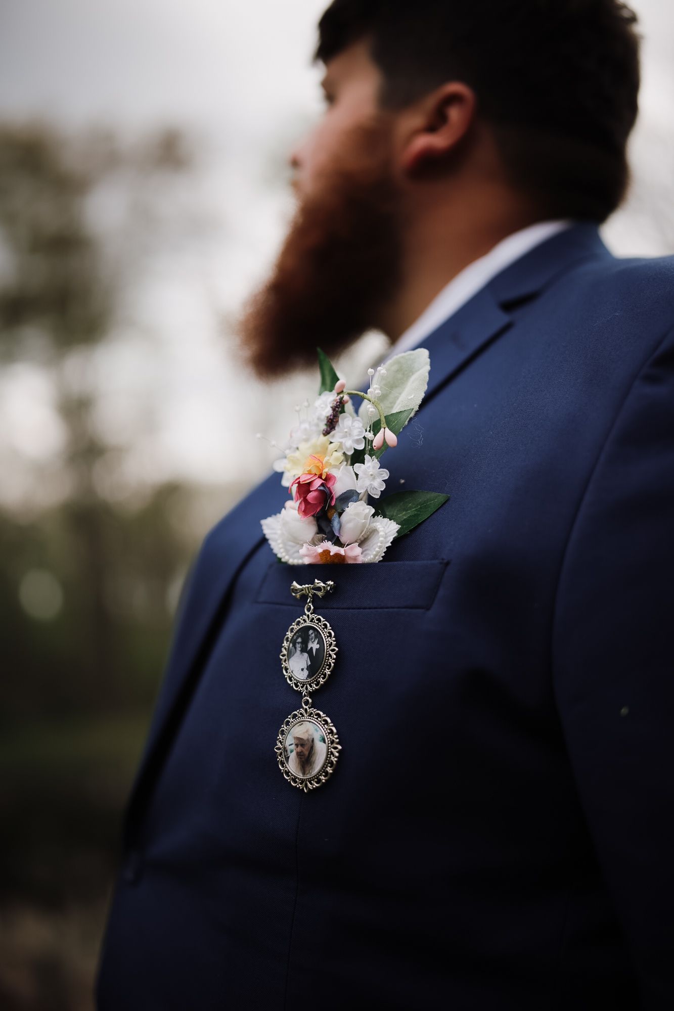Groom in navy suit with floral boutonniere and memorial locket charm, beard visible, outdoor wedding setting.