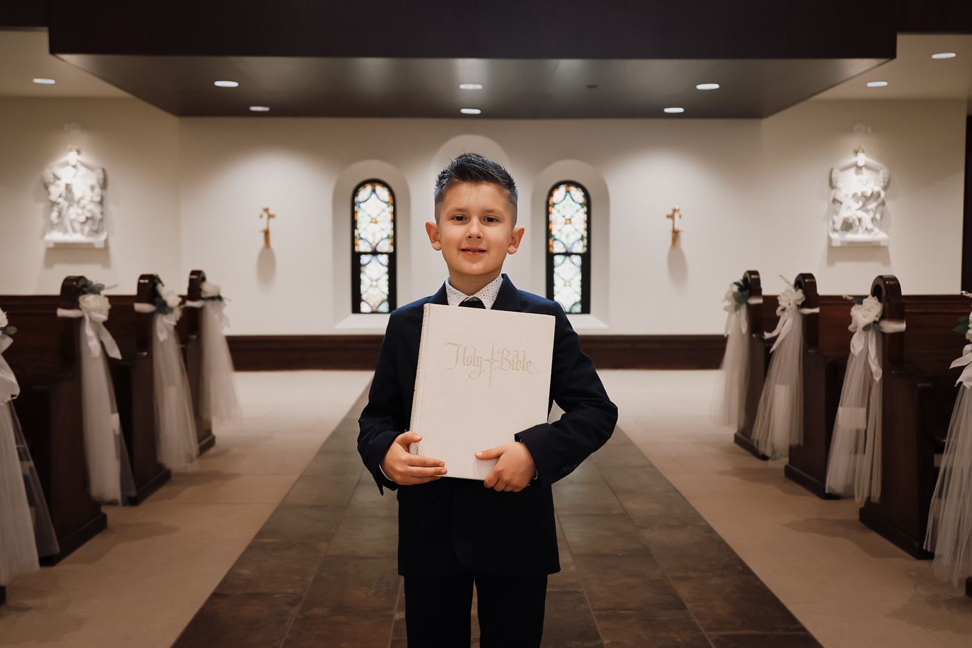 Boy in navy suit holding a white book in a decorated church aisle during First Communion ceremony.