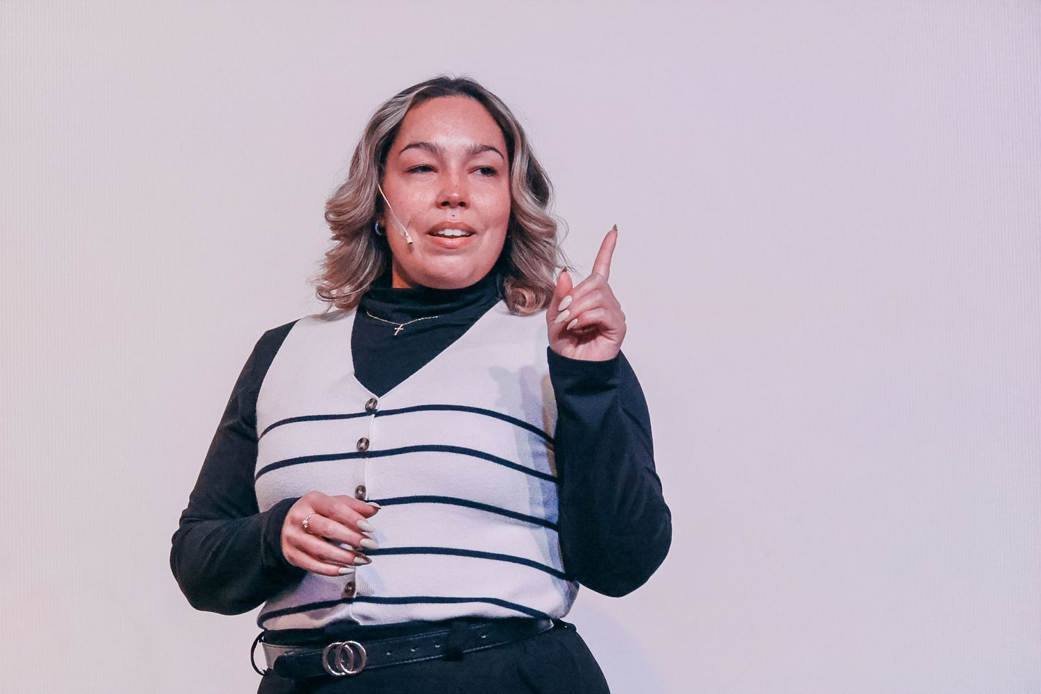 Woman in striped vest pointing finger up against light background, making a point gesture.