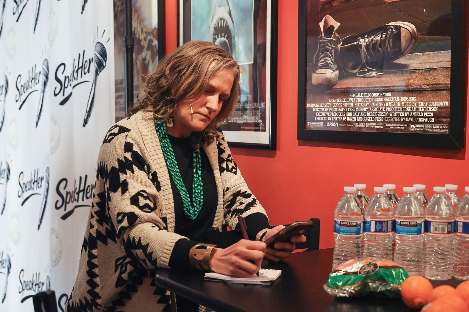 Woman with curly hair wearing a patterned cardigan and turquoise necklace using her phone at a signing event.