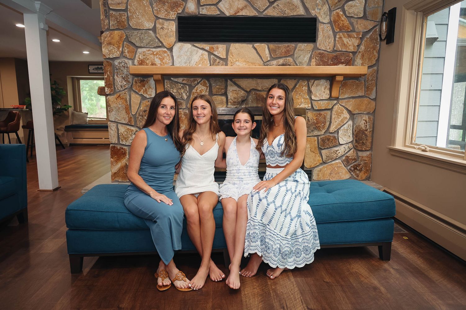 Four women smiling together on a teal sofa in front of a stone fireplace in a modern living room.