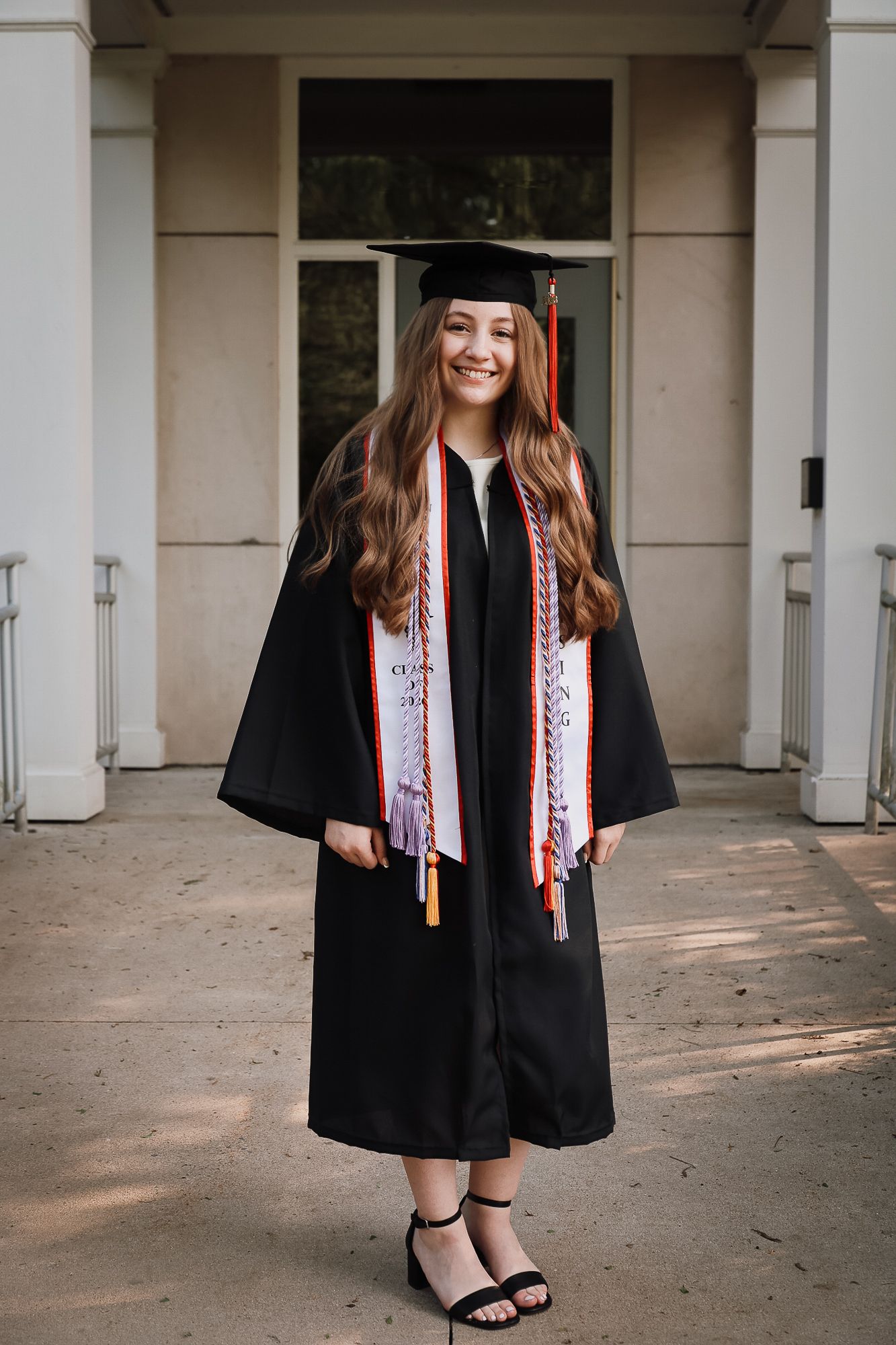 Smiling young woman in black graduation cap and gown with colorful stoles poses outdoors near building columns.