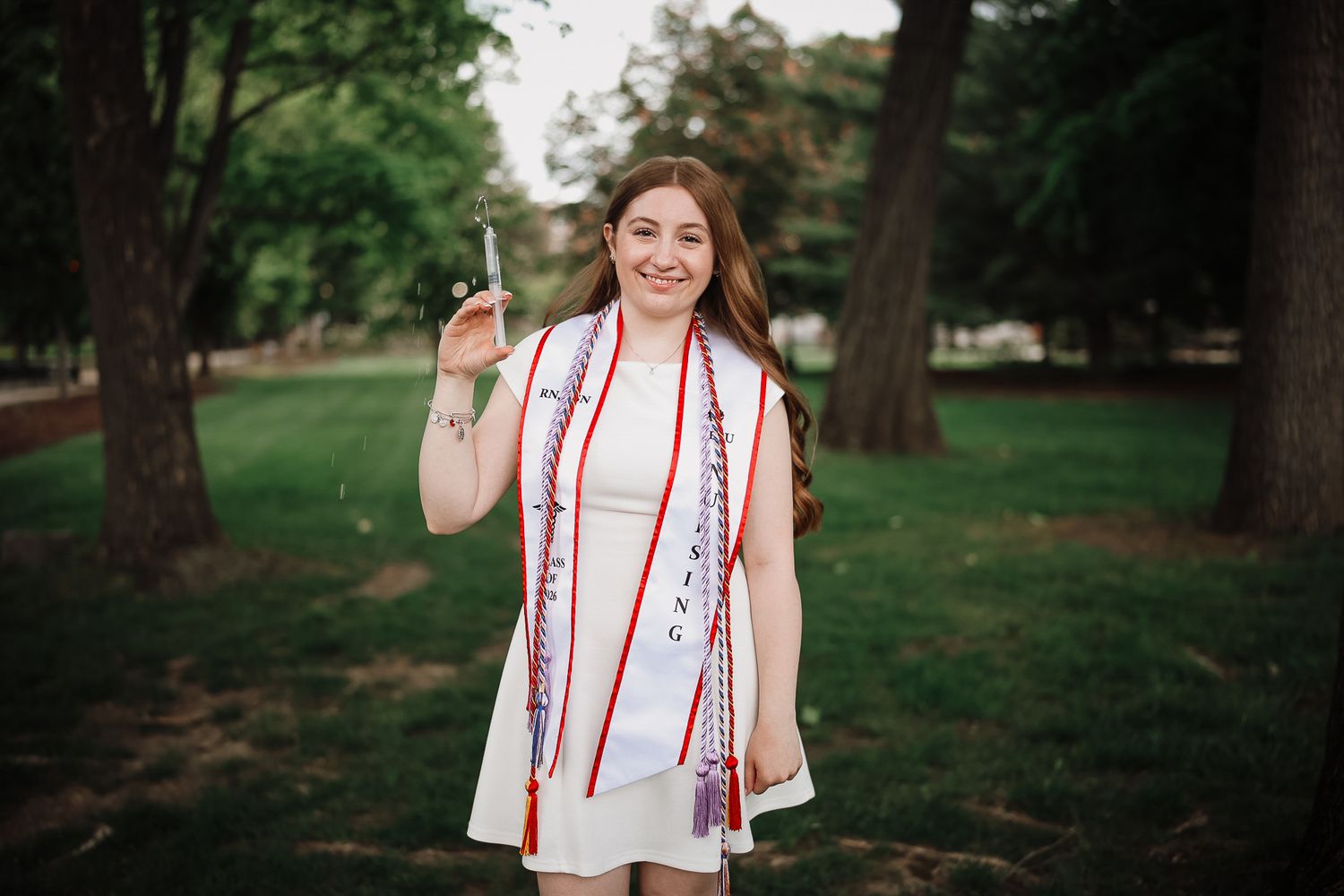 Young woman in white graduation gown holding diploma, wearing red sash and leis, smiling outdoors on campus.