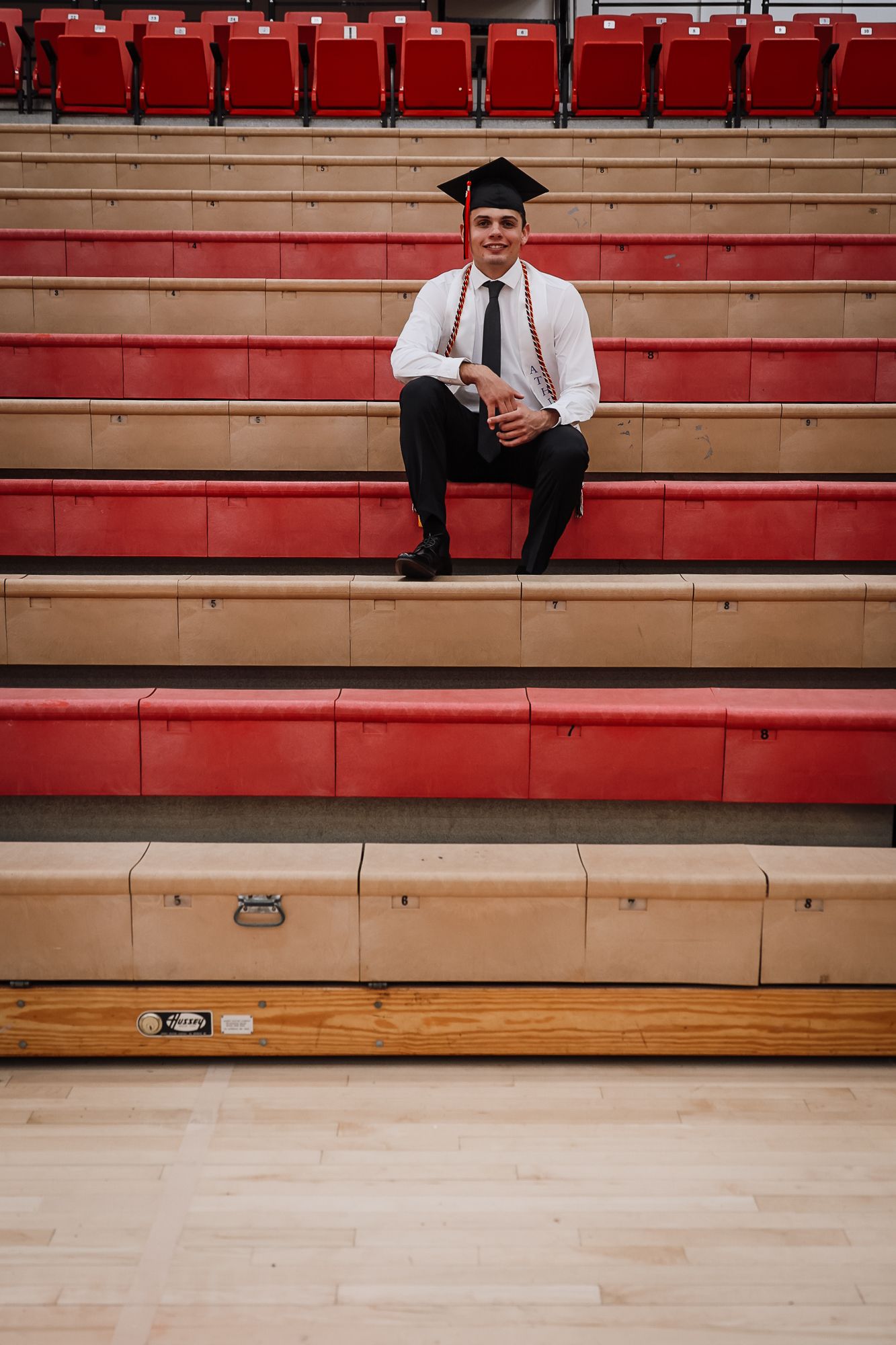 Graduate in cap and tie sitting on red bleacher steps in a gymnasium, smiling for a graduation photo.