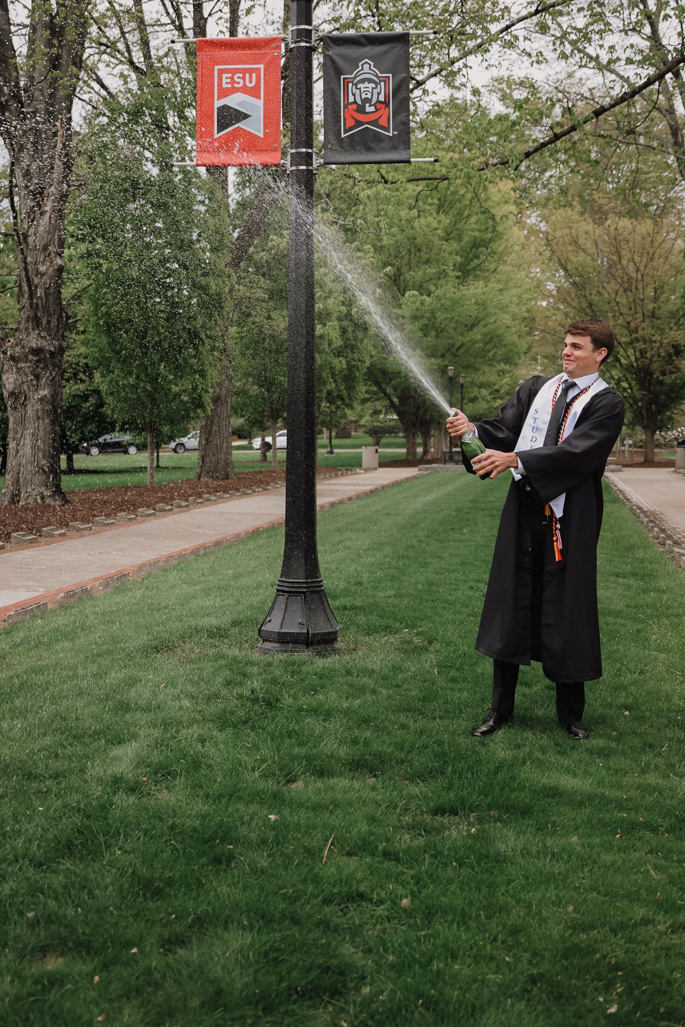 Graduate in cap and gown spraying champagne on ESU campus lawn during graduation celebration.